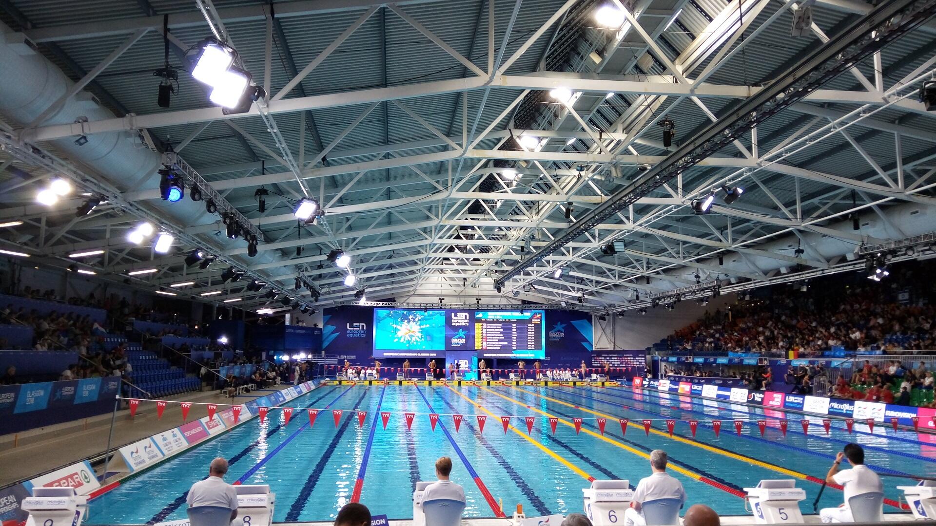 An indoor swimming pool with a large scoreboard at the hall's far end and spectators in the stands.
