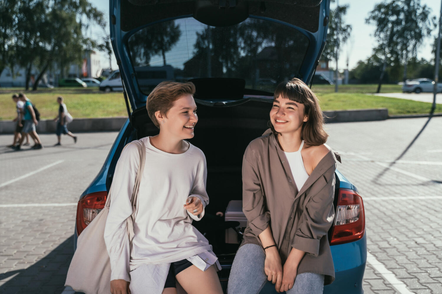 Two women sitting on the open trunk of a blue car, chatting outdoors on a sunny day in a parking area.