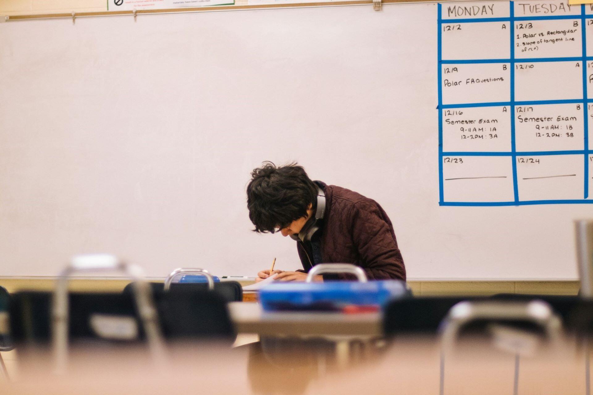 Student wearing headphones writing at a desk in a classroom with a wall calendar