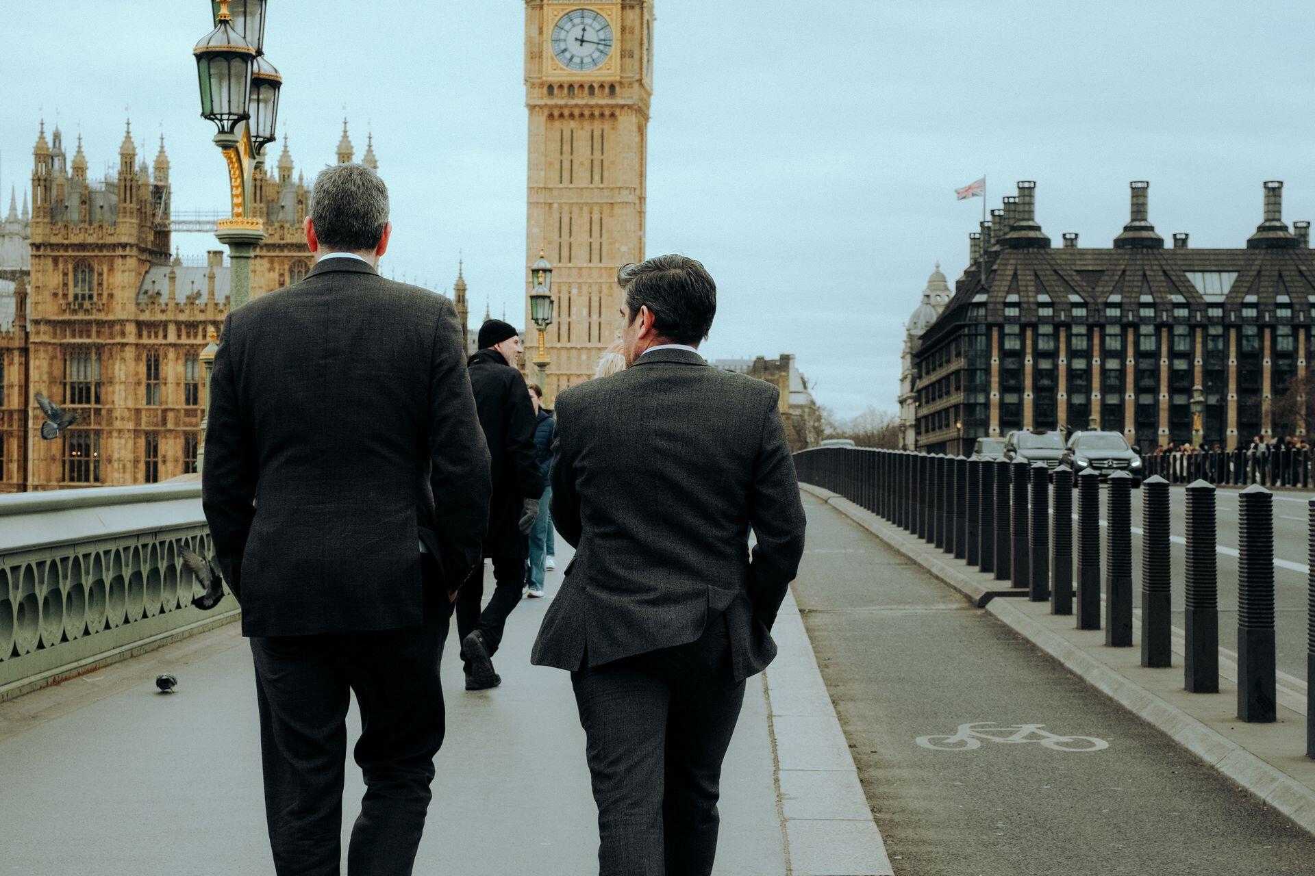 Two men in suits walking on a London bridge with Big Ben and historic buildings in the background on a cloudy day.