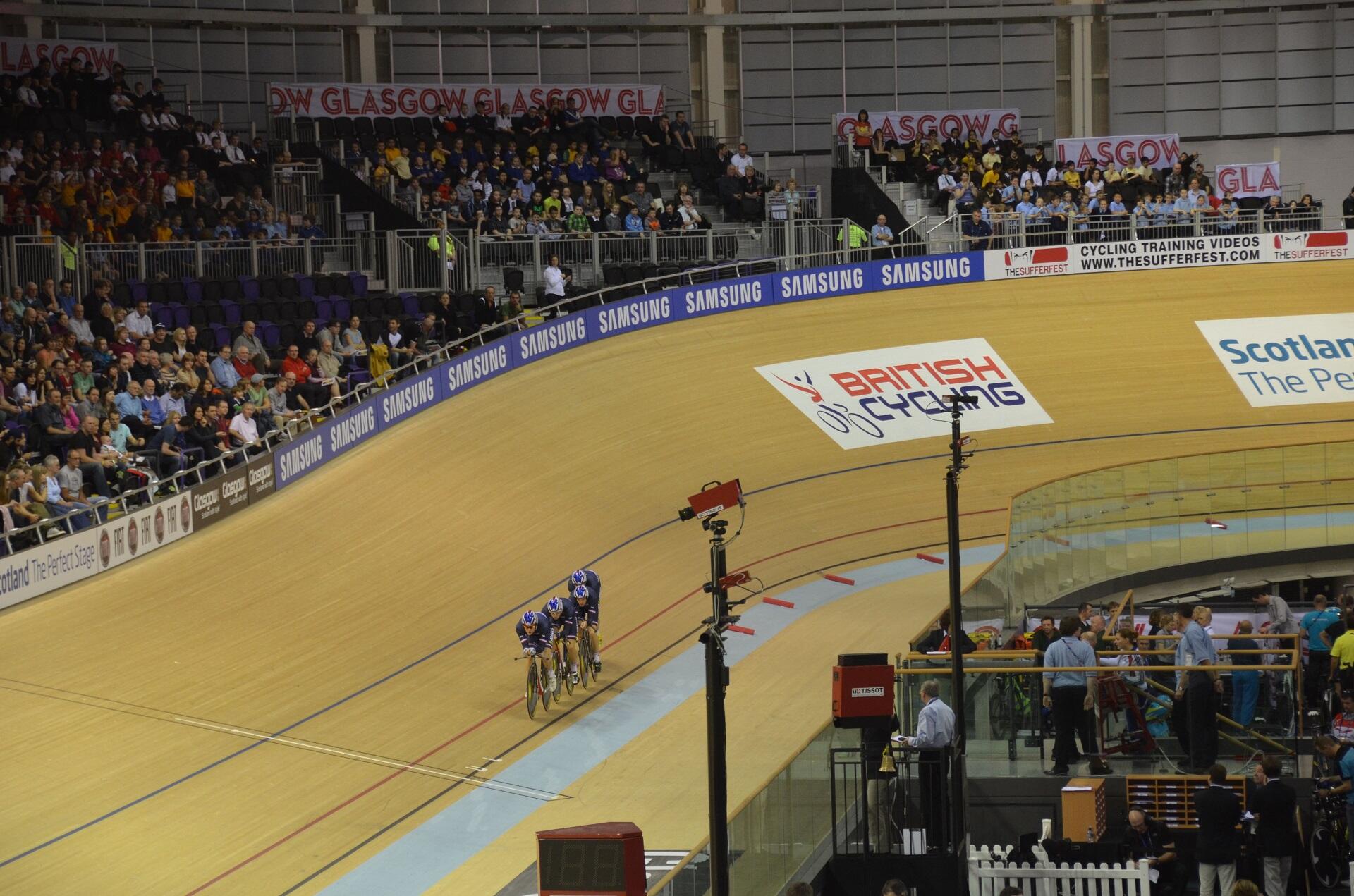 An indoor blond wood track with cyclists racing and spectators in the stands.