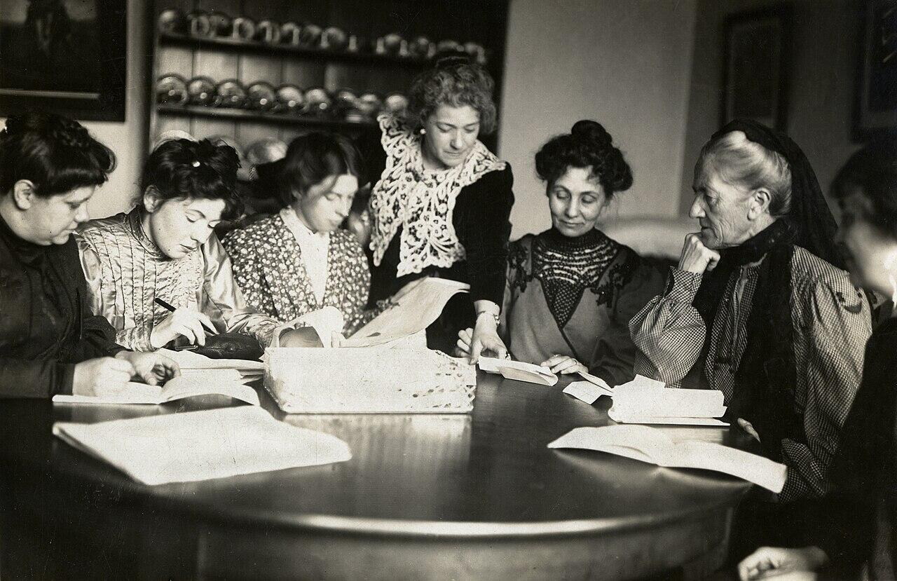 A group of women in historical dress are gathered around a round table, engaged in writing and reading documents.
