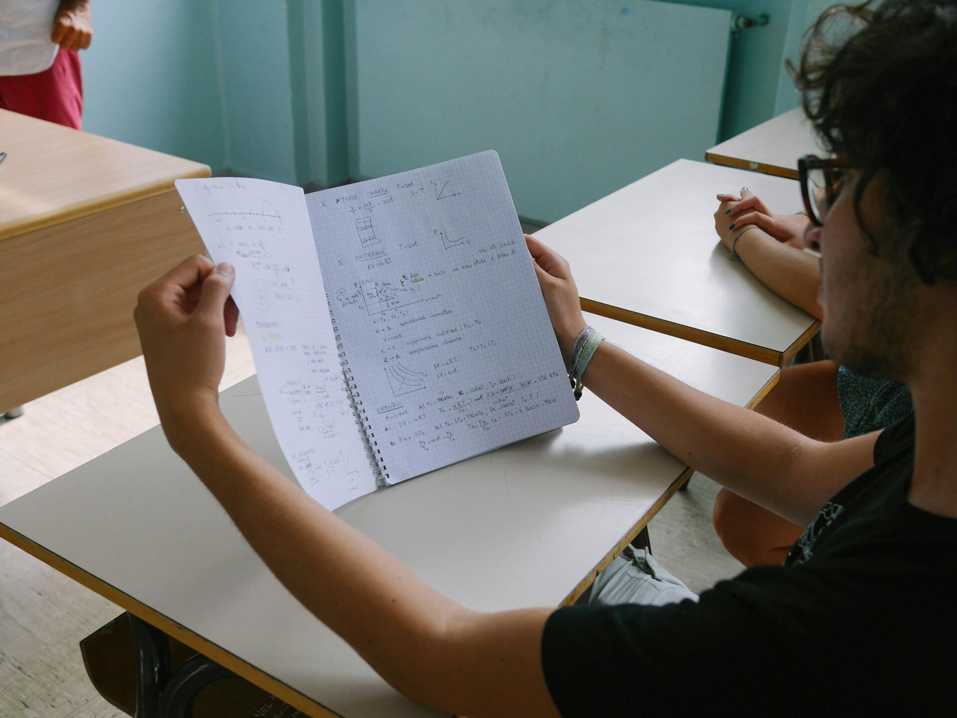 A person seated at a classroom desk looks over notes in a notebook while others are nearby.