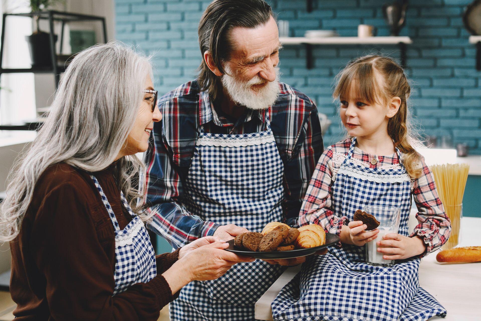 Grandparents and a granddaughter in matching aprons share baked goods and milk in a kitchen.