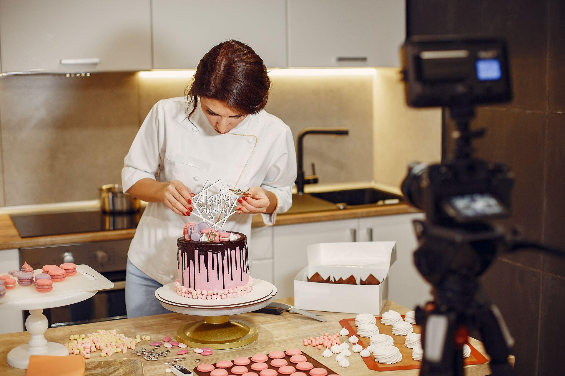 Baker decorating a cake while a camera records in a kitchen.