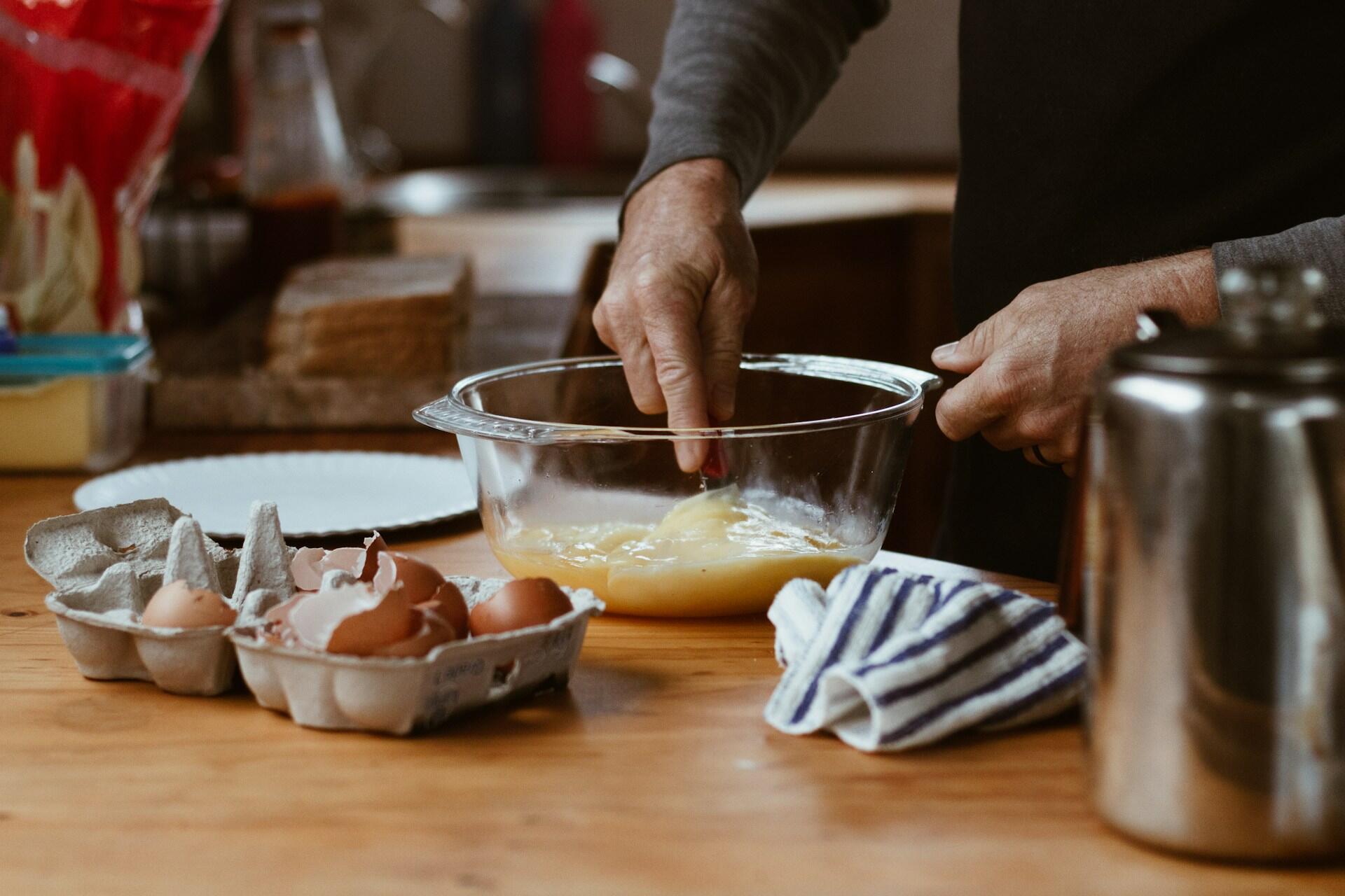 Hands whisking eggs in a glass bowl with an egg carton and shells on a wooden table.