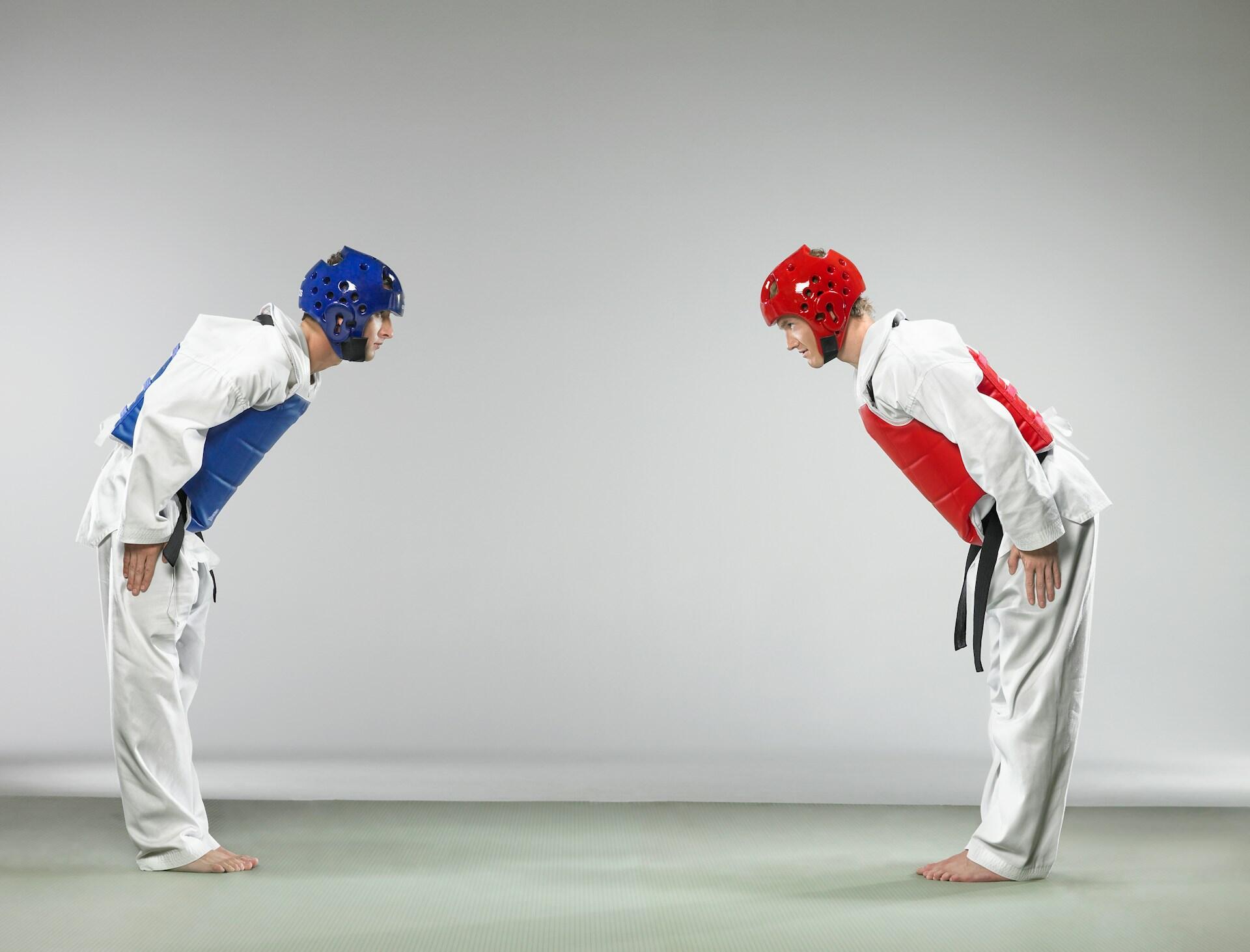 Two people in martial arts clothing and protective gear bow at one another.