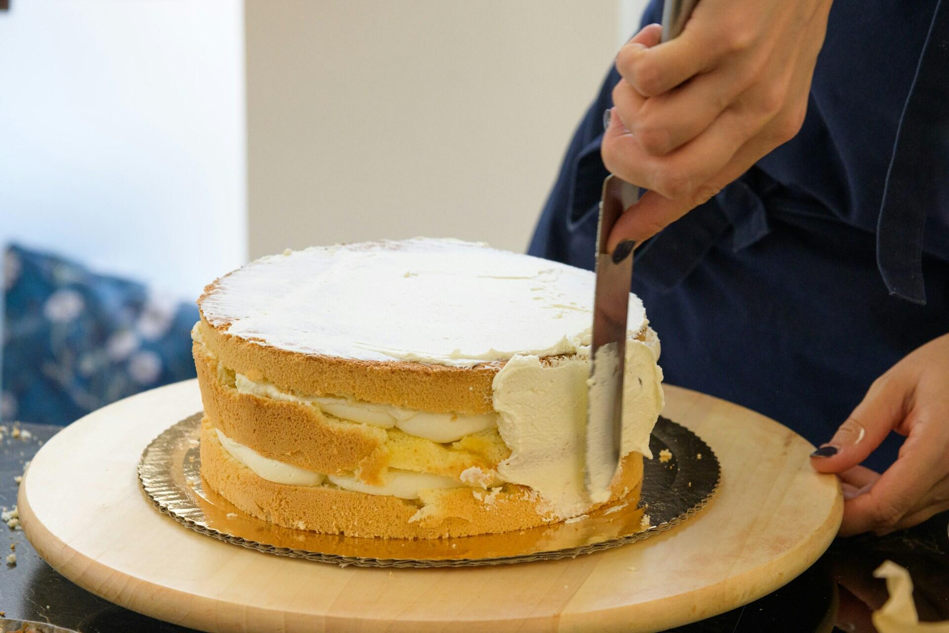 Hands frosting a multi-layered naked cake on a wooden cake stand with a metal spatula.