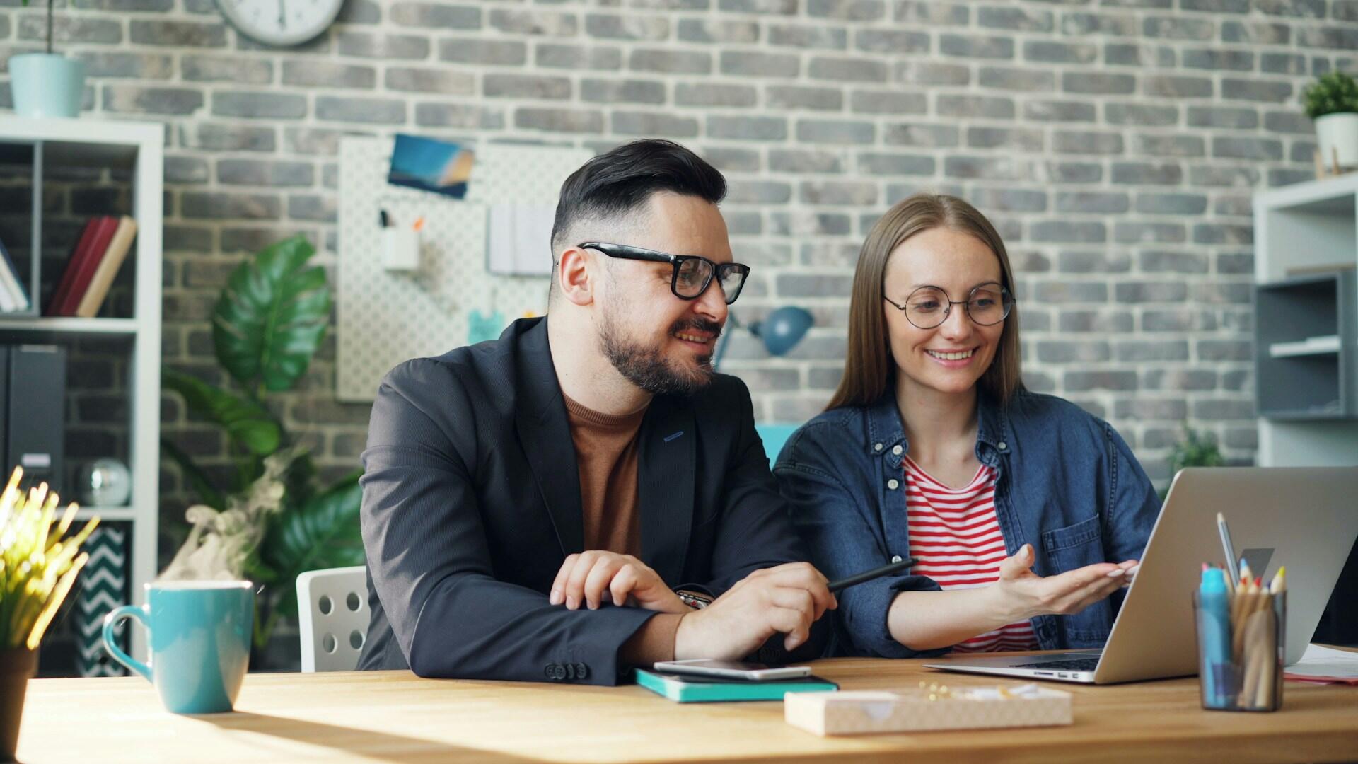 A man with glasses and beard, and a smiling woman wearing round glasses and a striped shirt, collaborate at a wooden desk while looking at a laptop in a modern office with exposed brick walls.