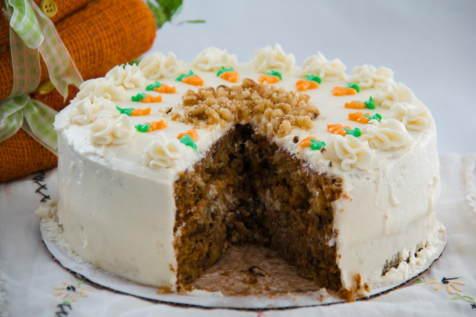 A slice has been cut from a round carrot cake with cream cheese frosting, topped with walnuts and decorated with small frosting carrots and white swirls.