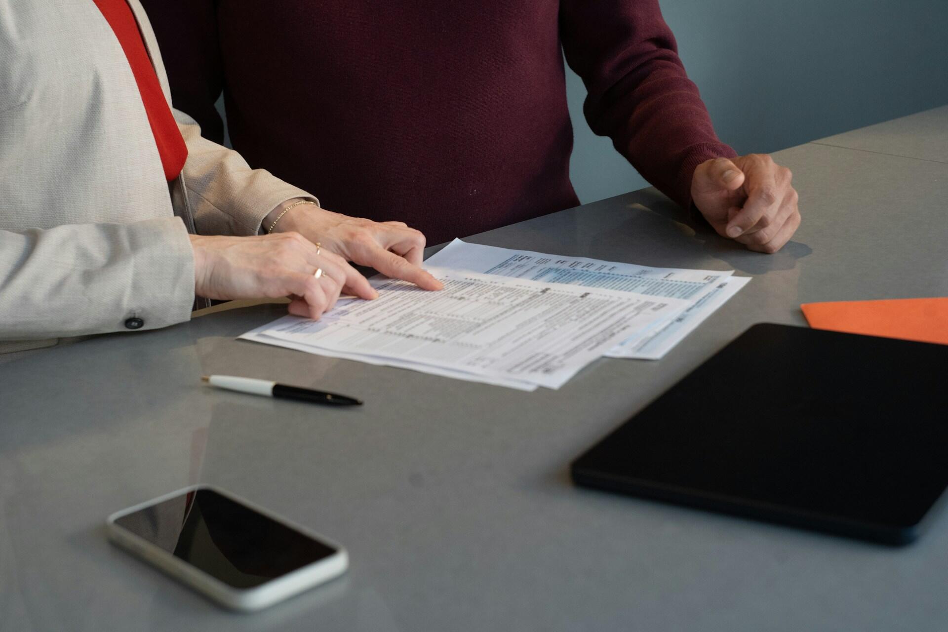 Two people review paperwork at a table, with a pen, smartphone, and black folder nearby.