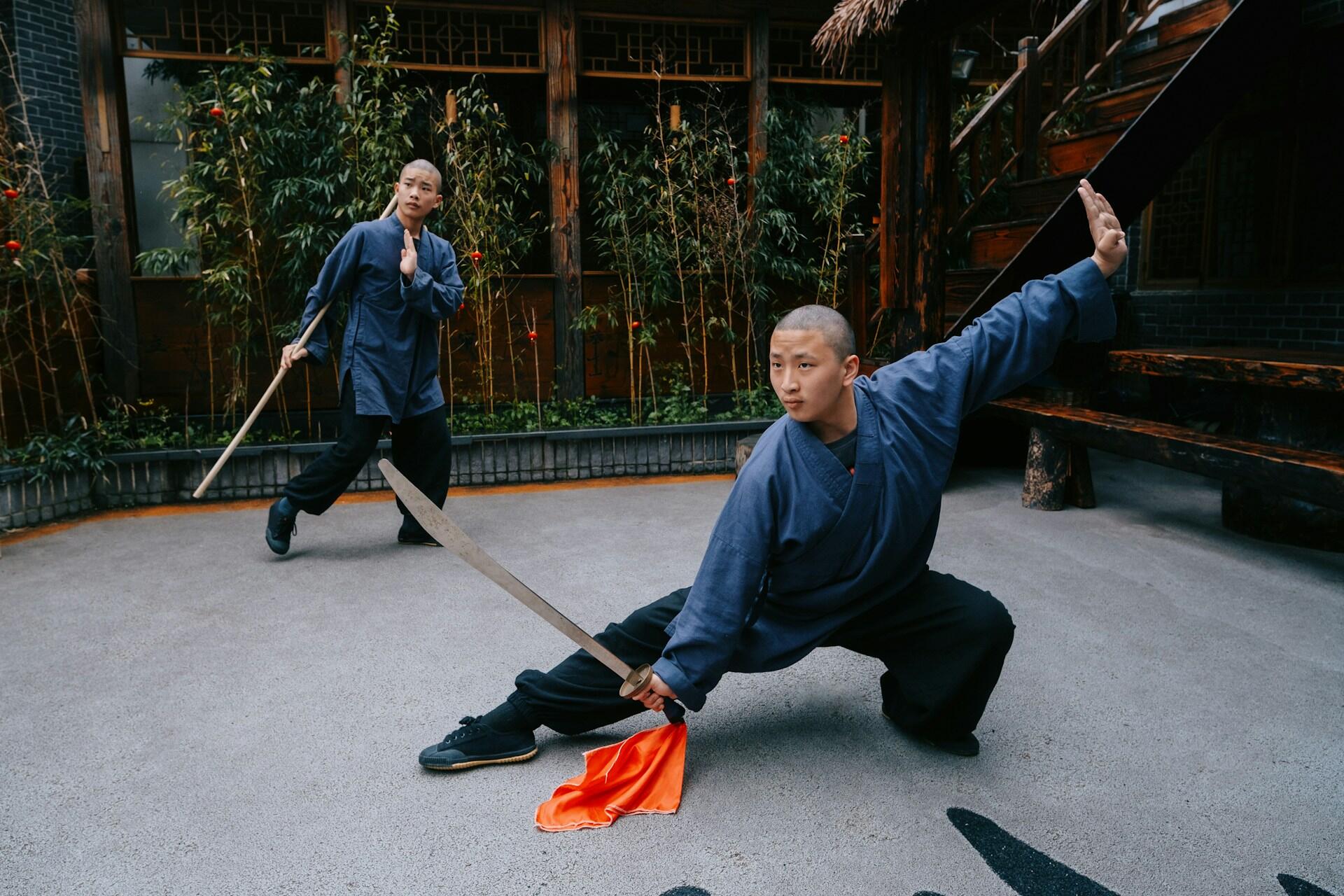 A man wearing a blue gi holds a bo and strikes a pose while another one looks on.