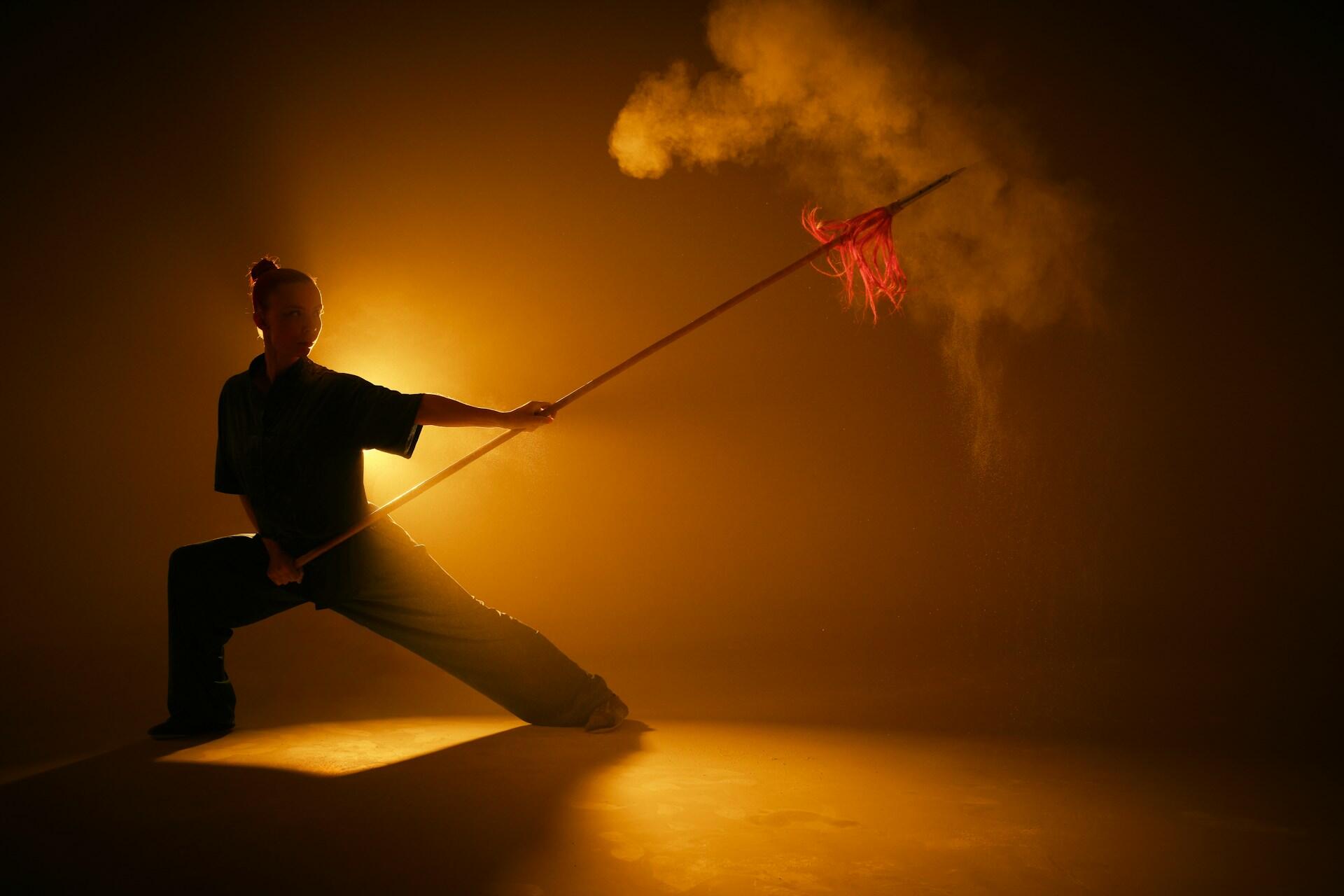 A person wearing martial arts clothing holds a spear while standing in a defence position against a lit-up background.