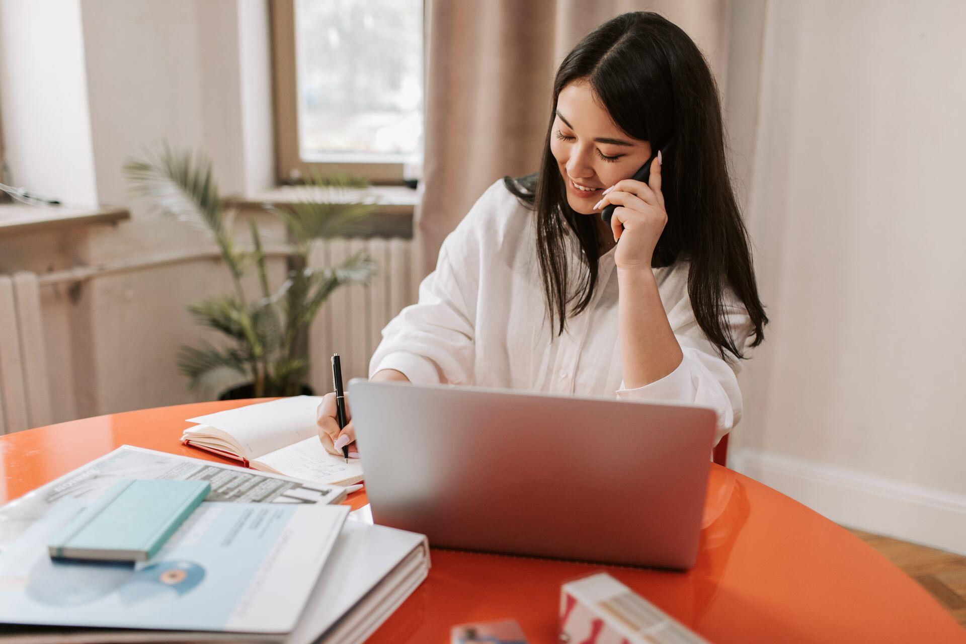 A young woman with long dark hair sits at an orange desk, smiling while talking on her phone and taking notes in a notebook next to her laptop.