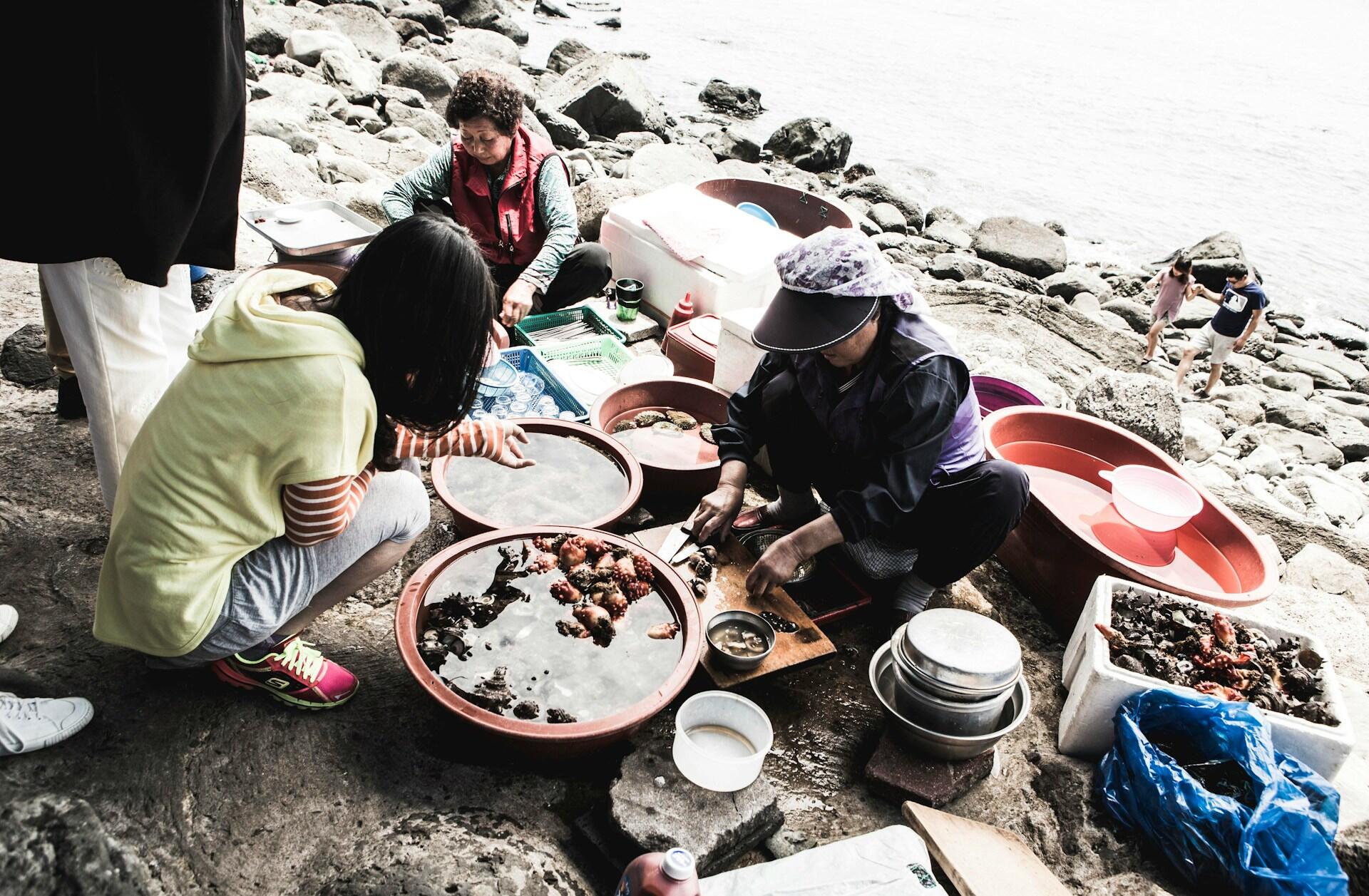 Two older women sit on a sandy beach sorting mussels while a young girl looks on.