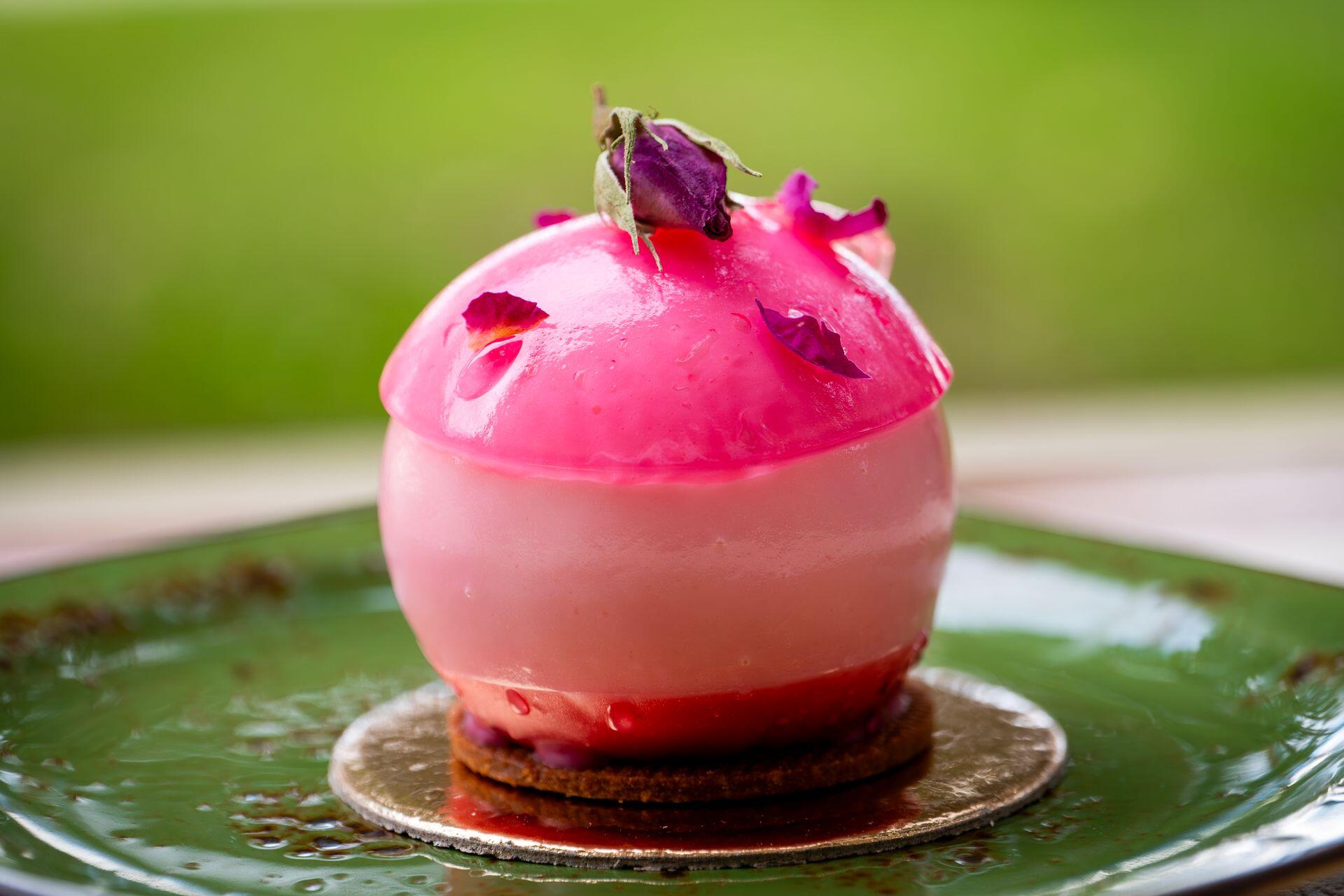 A pink, layered mousse dessert topped with rose petals and a rosebud rests on a green plate.