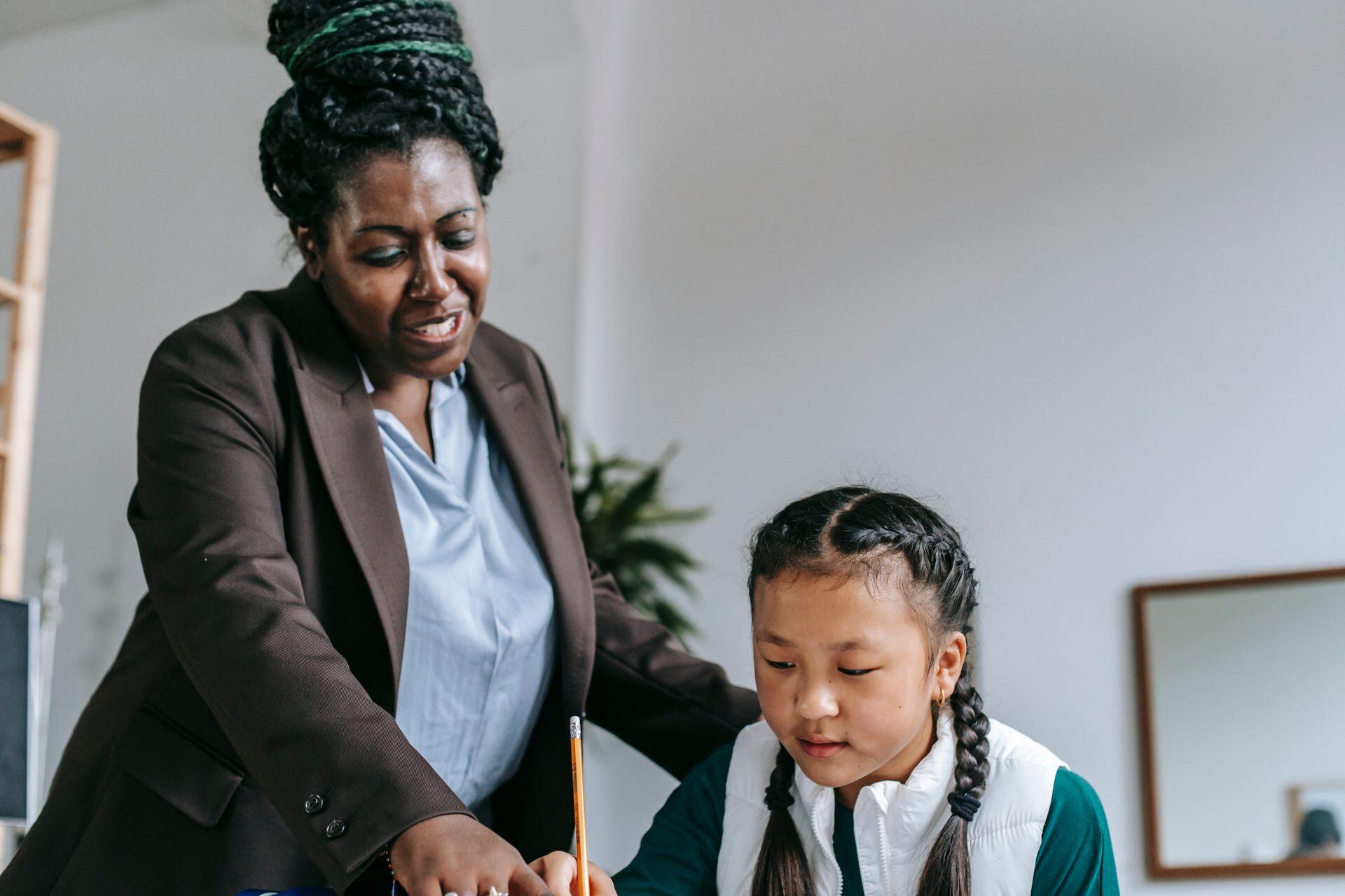 A Black woman in a brown suit guides a young Asian girl with braided hair as she writes with a pencil.