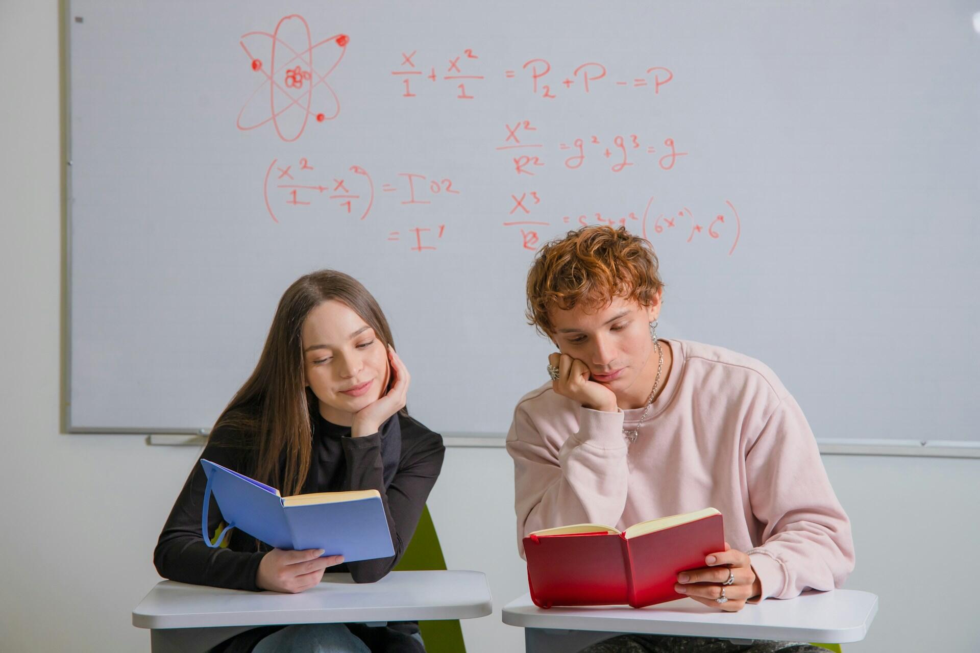 Two people sit side-by-side, one with a blue-covered book and the other with a red-covered one, looking bored.