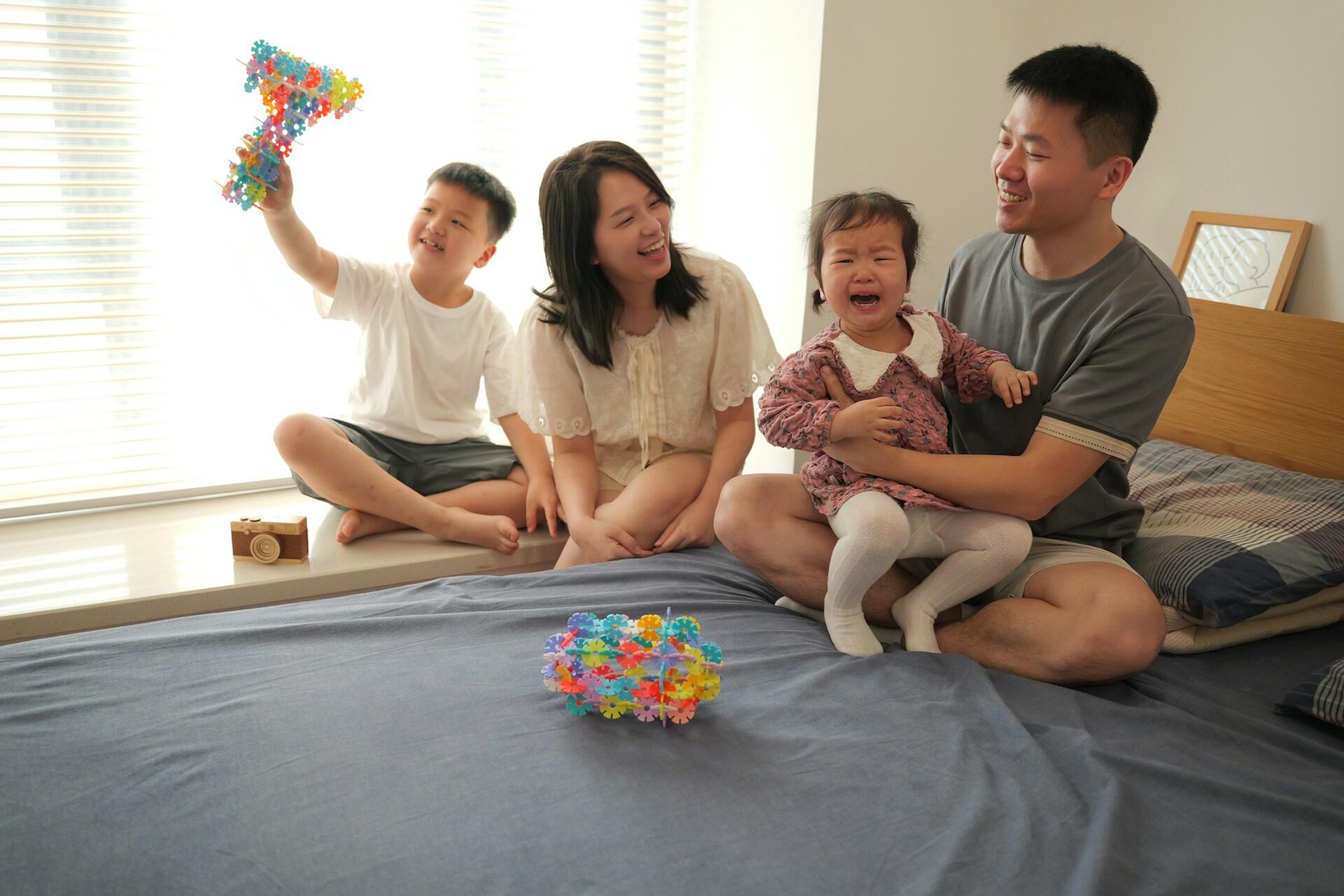 A family of mother, father, older brother and younger sister sit on a bed. The first three are happy but the sister is crying.
