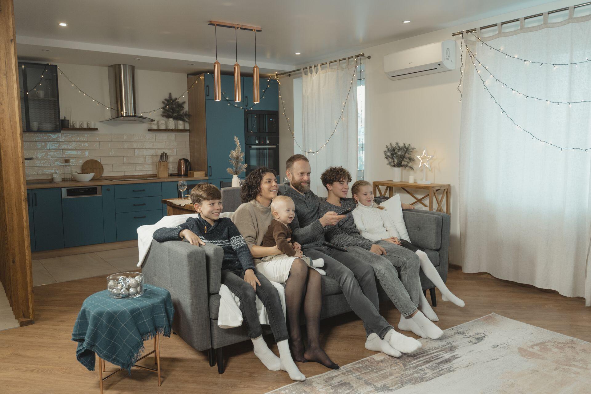 A family of six, including a baby, is watching TV together on a couch in a living room decorated with string lights.