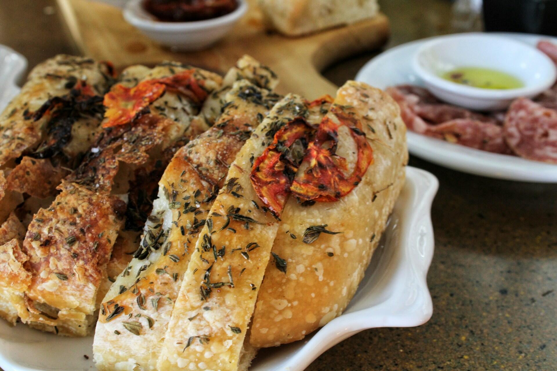 Crusty bread topped with tomatoes and herbs served in a white dish with salami and olive oil in the background.