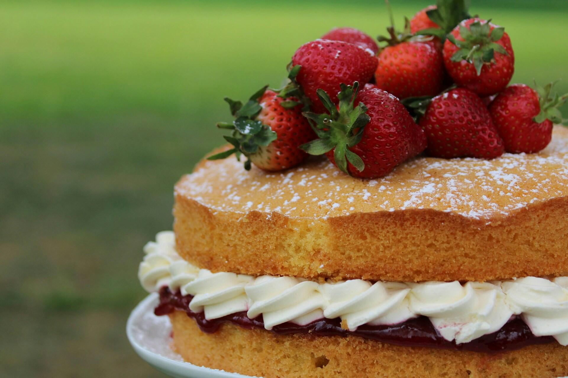 A Victoria sponge cake filled with cream and jam, topped with fresh strawberries and dusted with powdered sugar, sits outdoors on a plate.
