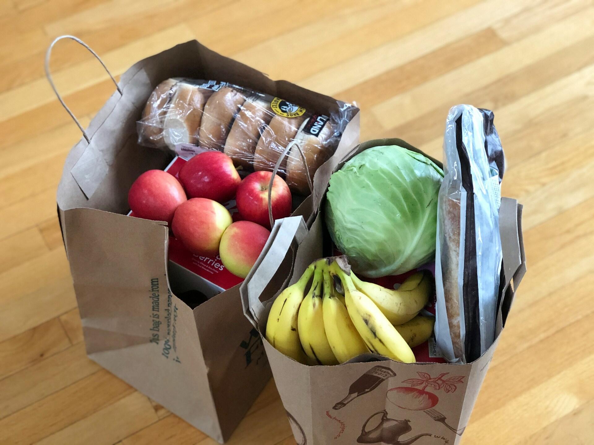 Two paper grocery bags filled with fresh produce, including red apples, bananas, a head of cabbage, and packages of bagels and bread, sit on a wooden floor.