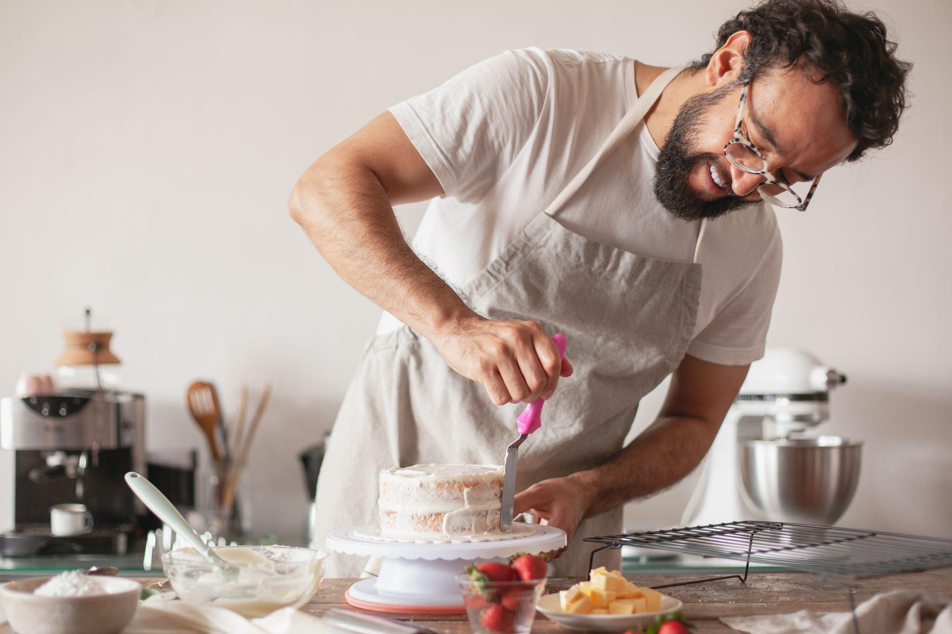 Man wearing an apron frosts a small white cake with a pink spatula.