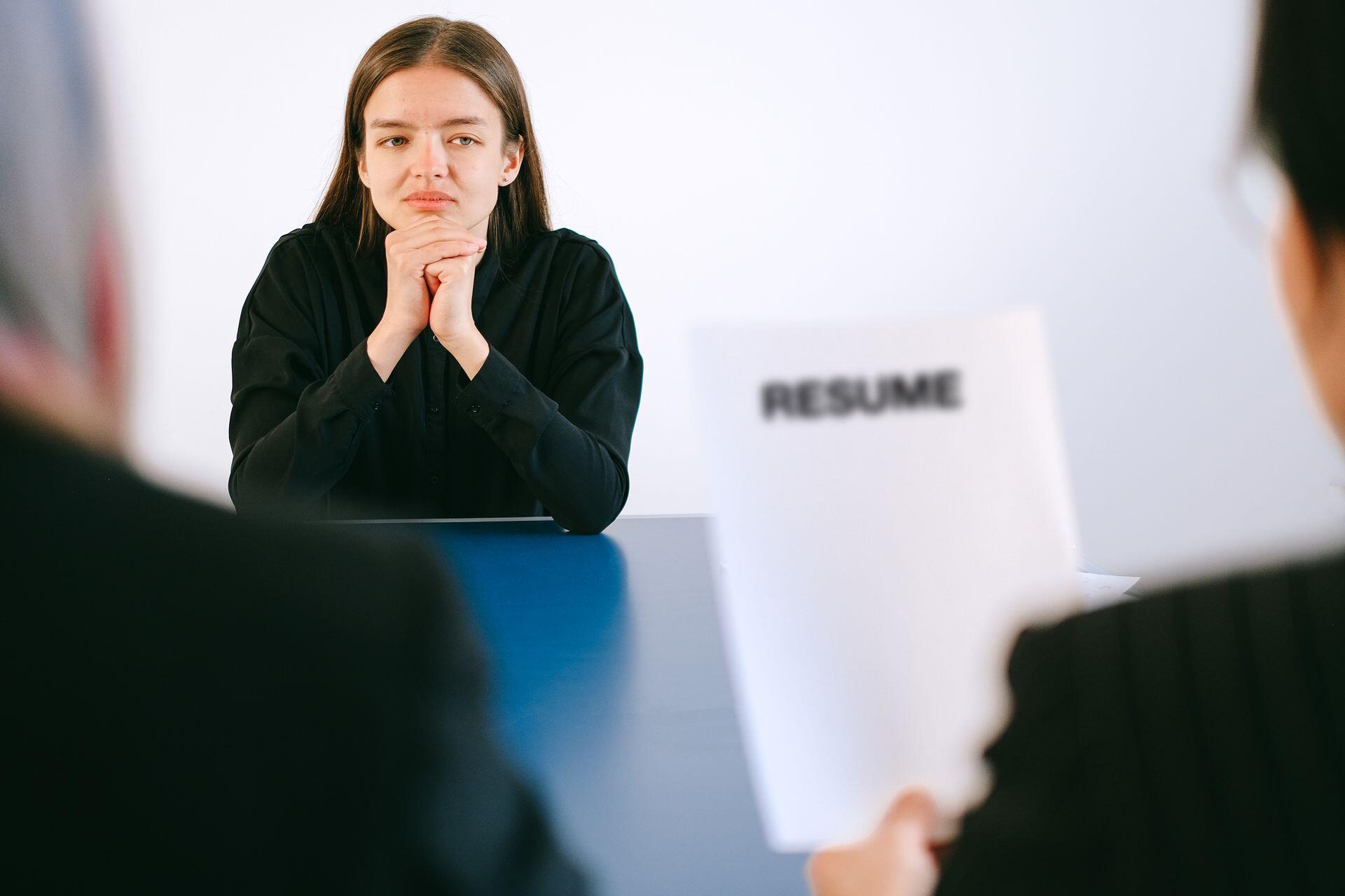 A serious job candidate sits with clasped hands during an interview while interviewers hold a résumé in the foreground.