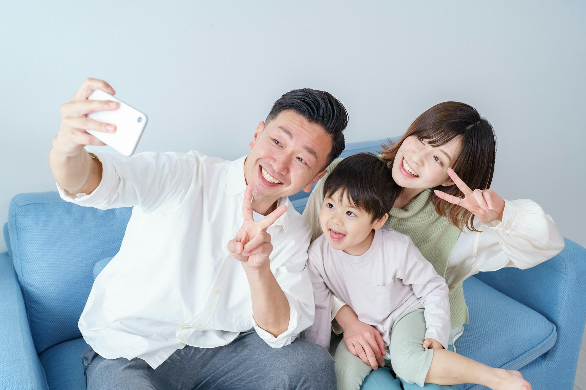 A young couple sits on a couch with a toddler between them posing for a selfie.
