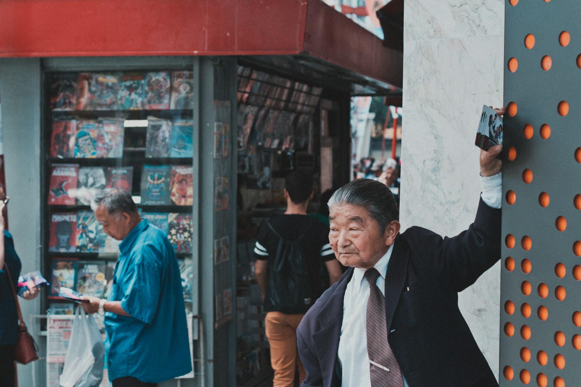A street scene with a man in a suit and tie in the foreground, another dressed causally to the left, and several youths lined up at a kiosk selling magazines.