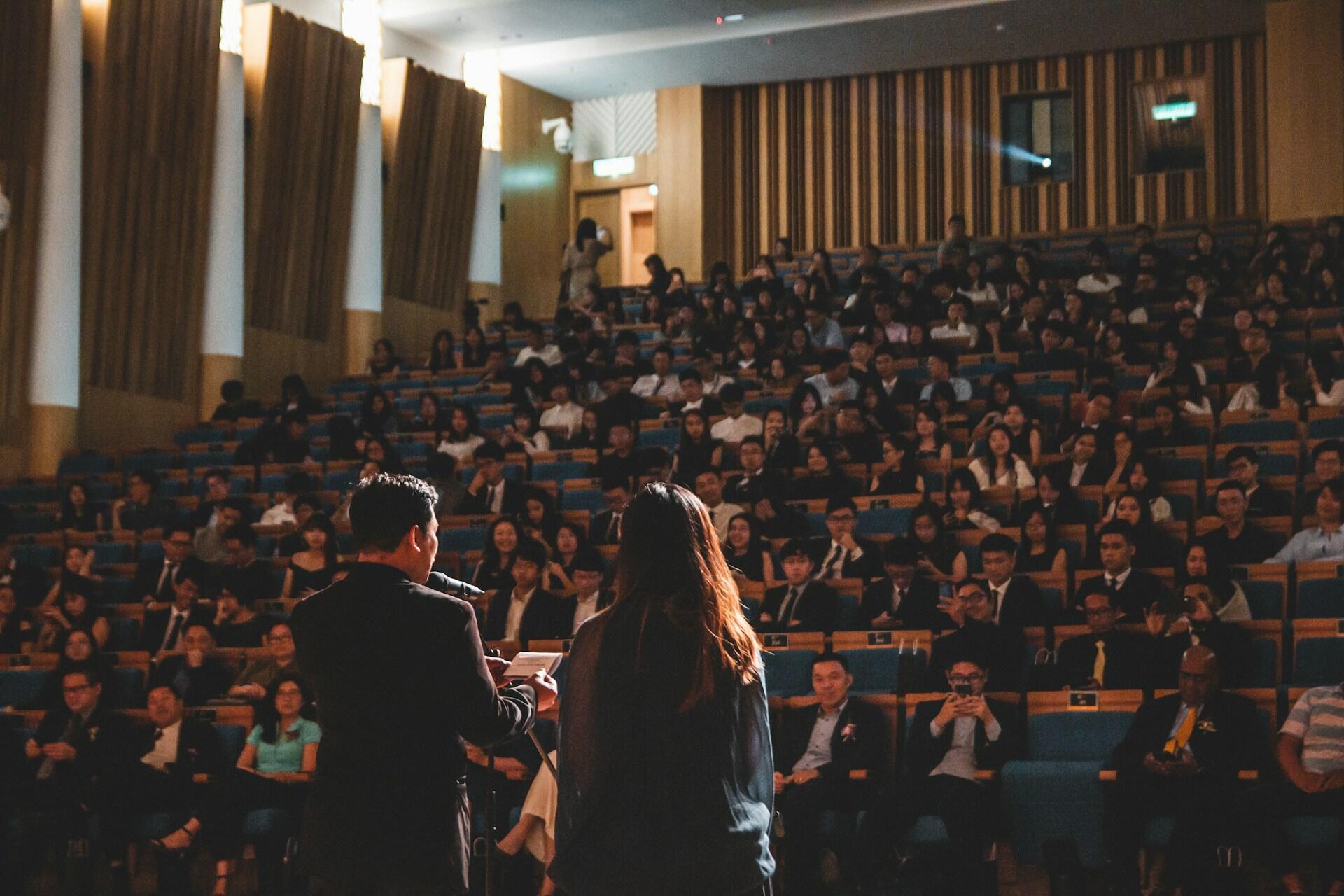 A man and woman in business clothing stand on stage addressing a large audience in an auditorium.