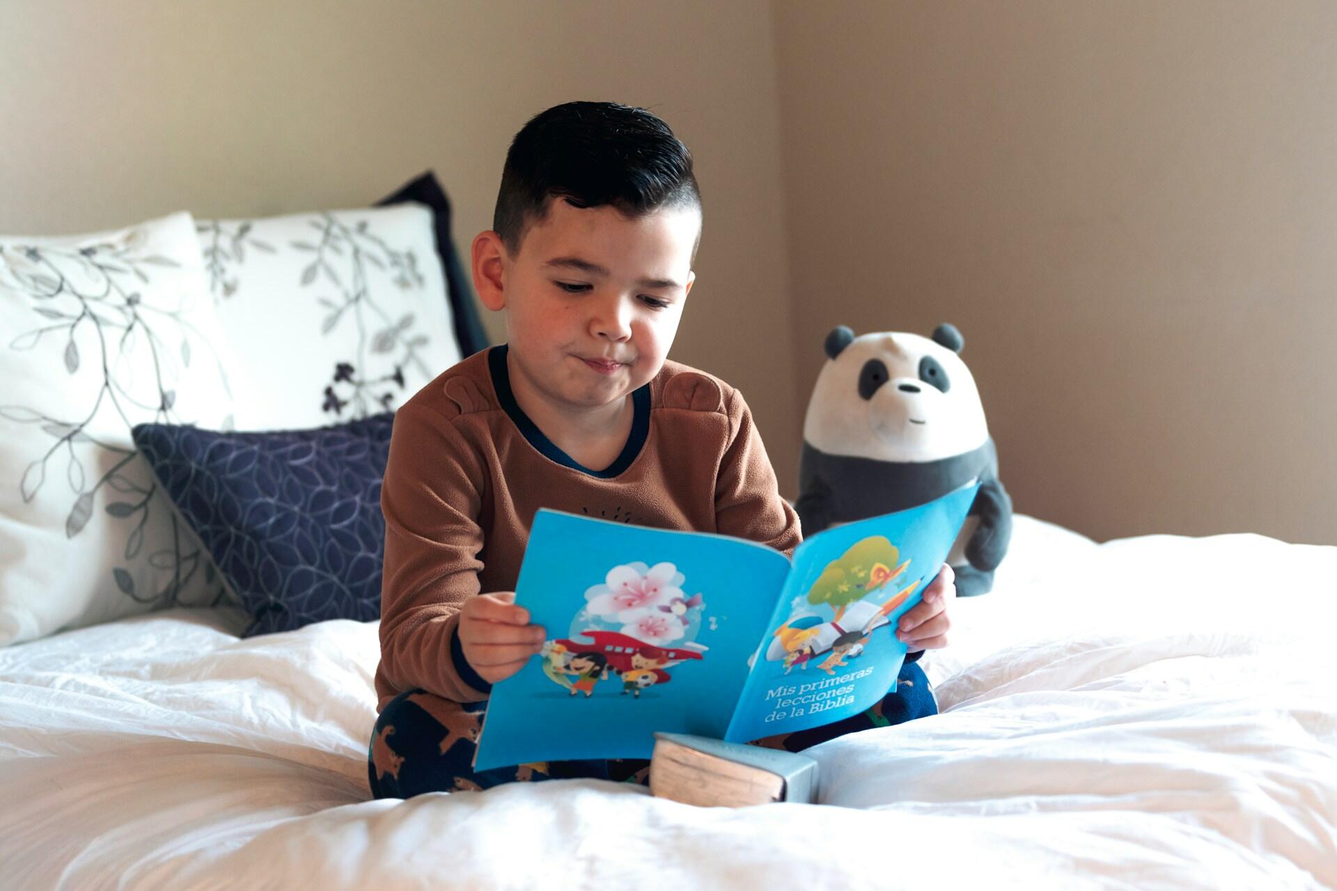 Young boy reading a book while he sits on bed, stuffed panda in background