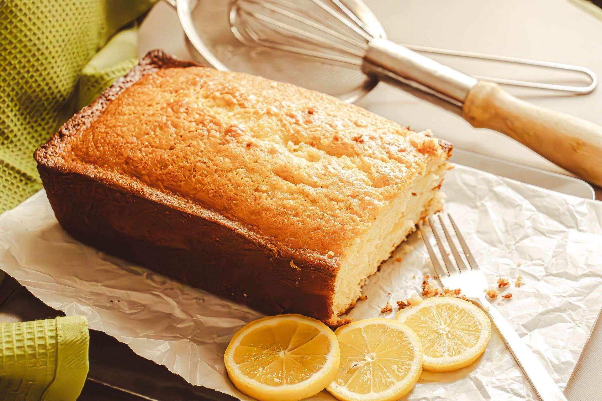 Golden brown loaf of lemon cake with sliced lemons and a whisk in the background.