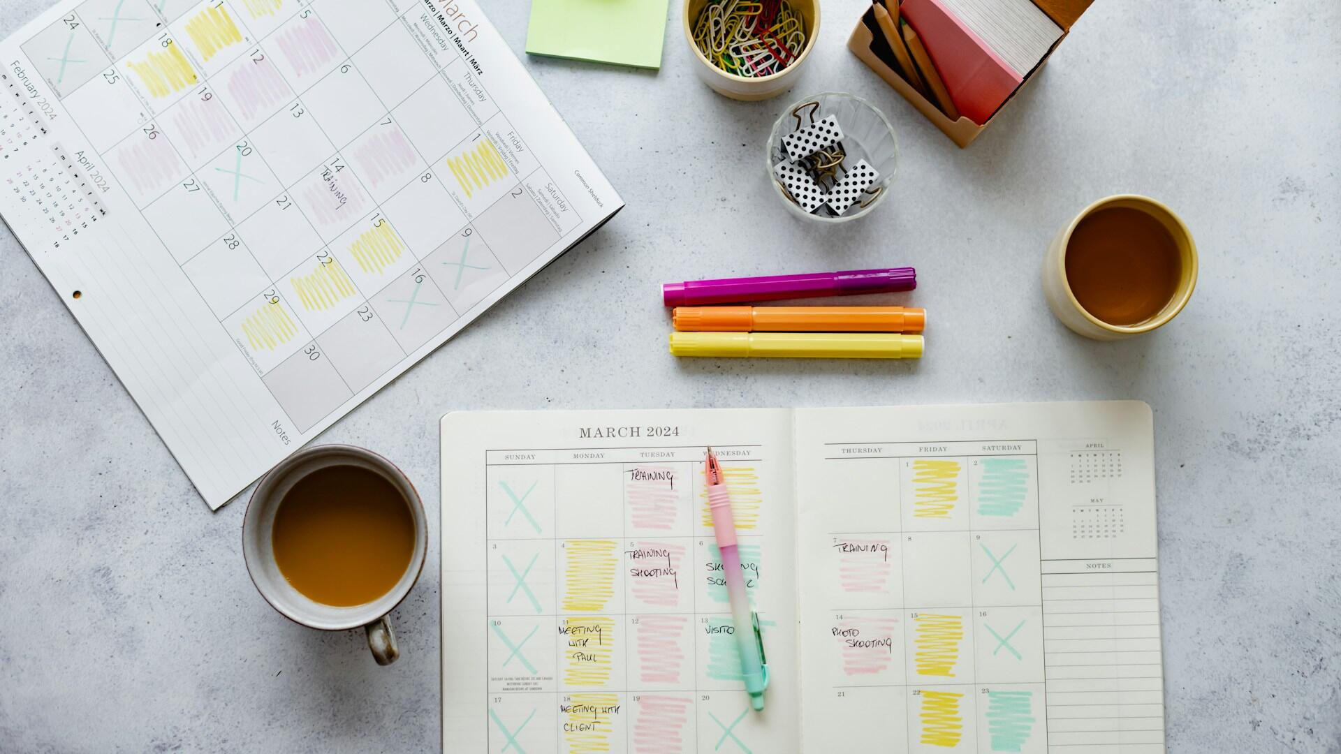 An open planner with a pen resting on it, with highlighters, a coffee cup, and other study necessities nearby.