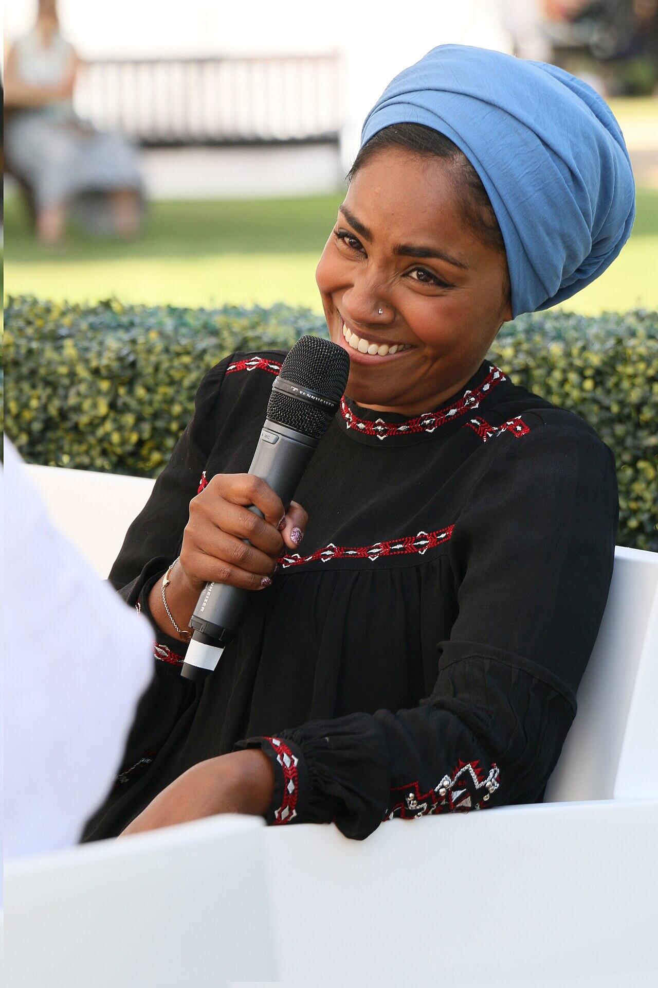 A Black woman wearing a blue headscarf and a black dress with red embroidery smiles while holding a microphone.