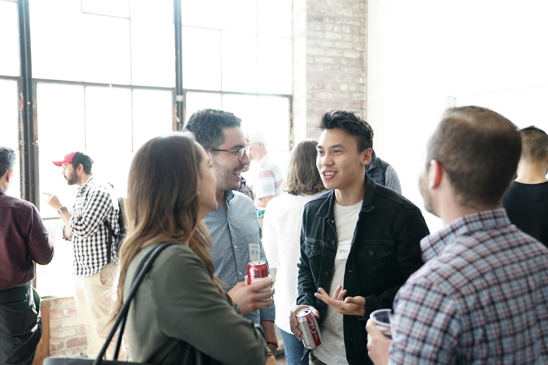 Group of diverse people mingling and talking at an indoor event, some holding drinks.
