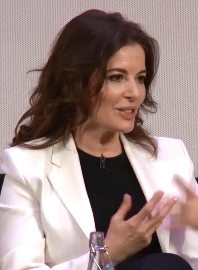 A woman with brown hair wearing a white blazer over a black shirt gestures with her hands while speaking.