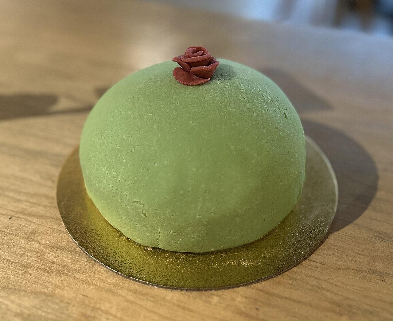 A green marzipan cake decorated with a red marzipan rose sits on a gold foil base on a wooden table.