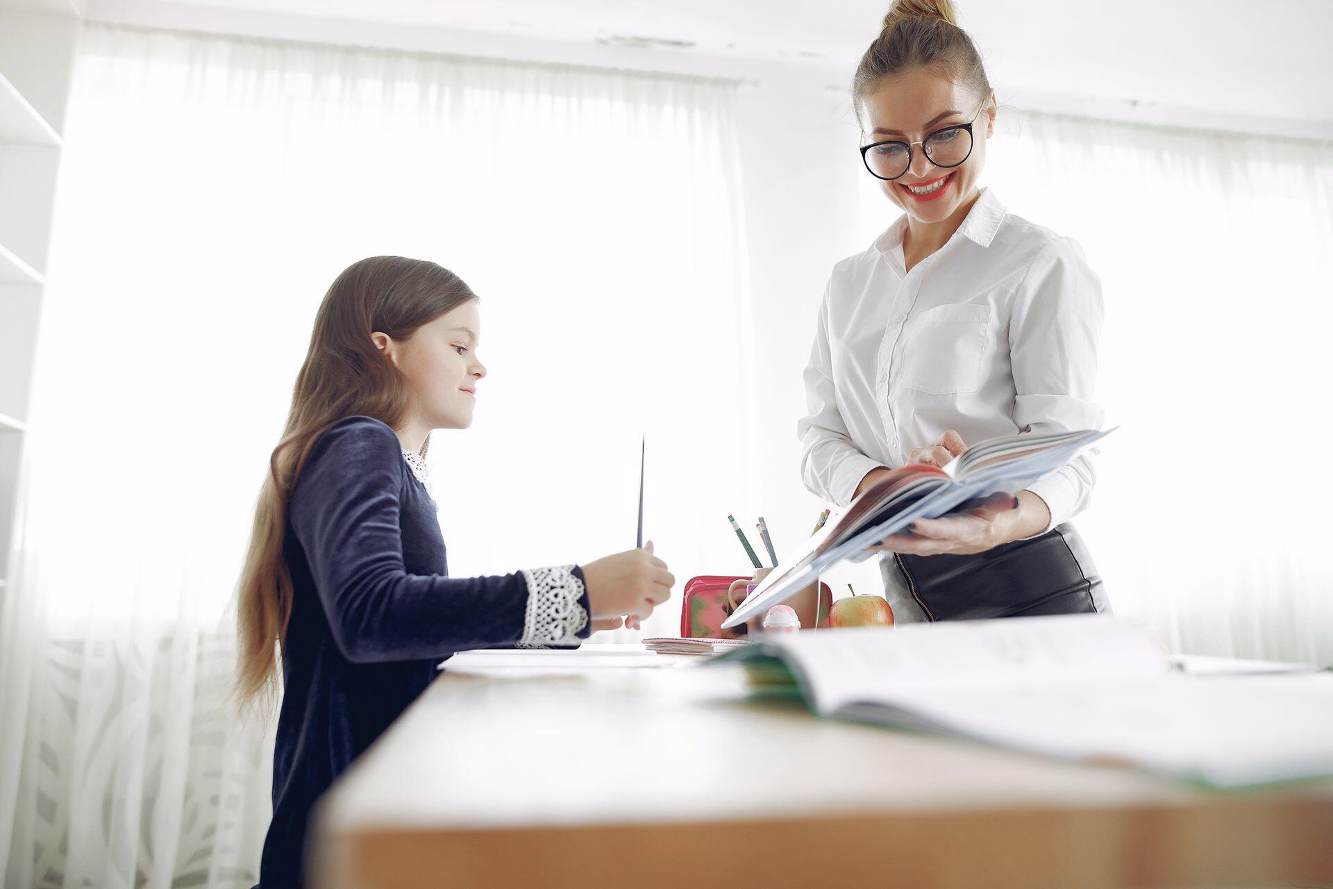 A teacher smiles as she holds a book while a student sits at a desk holding a pencil.