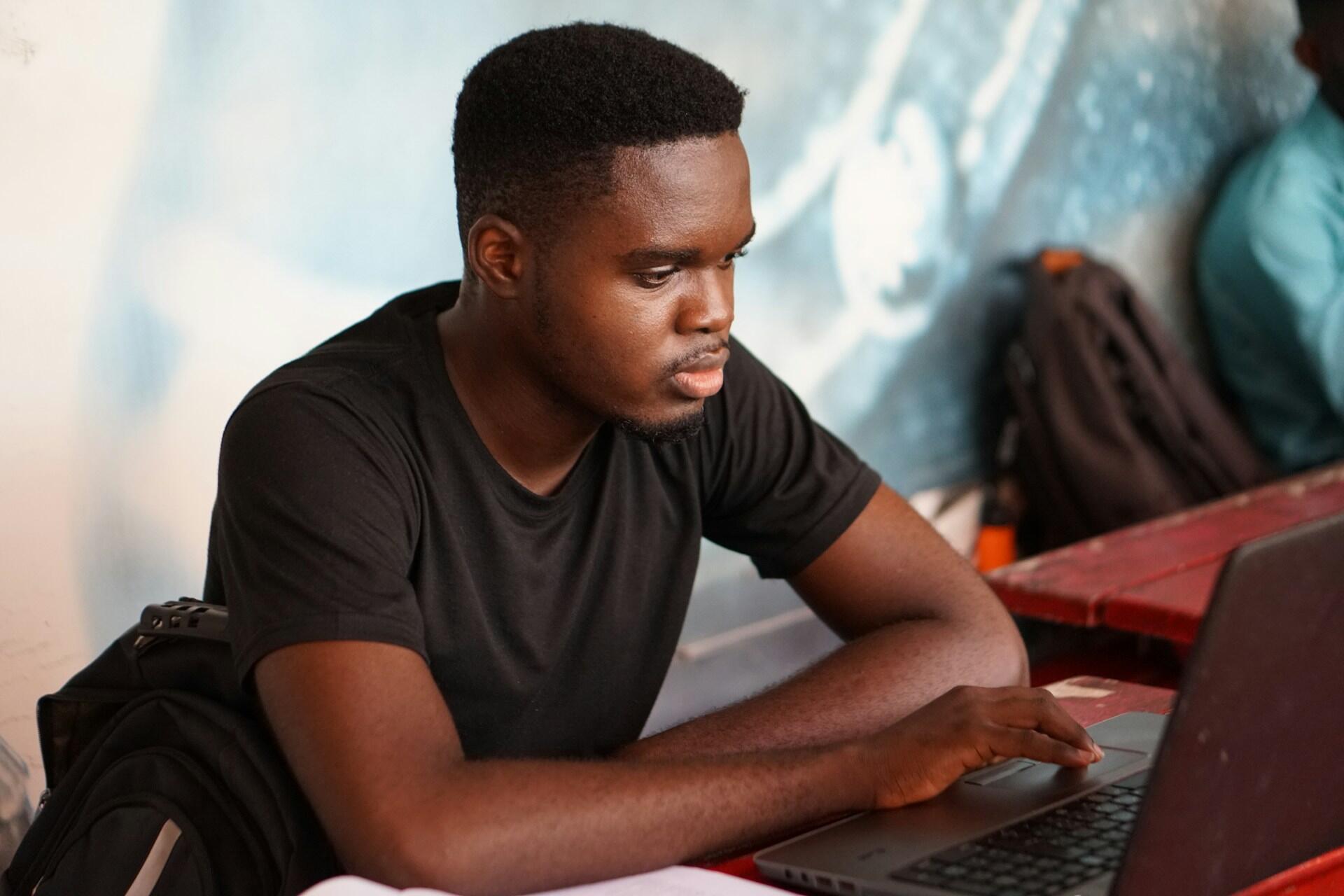 A young man wearing a black tee-shirt sits at a table working a laptop computer with his right hand.