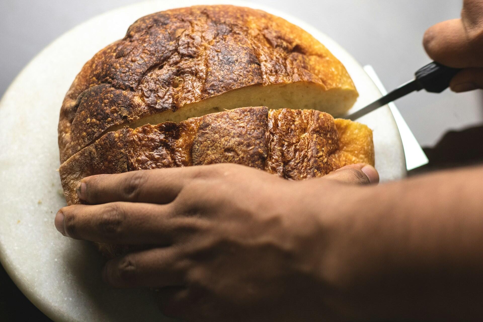 A hand slices a rustic loaf of bread on a marble cutting board.