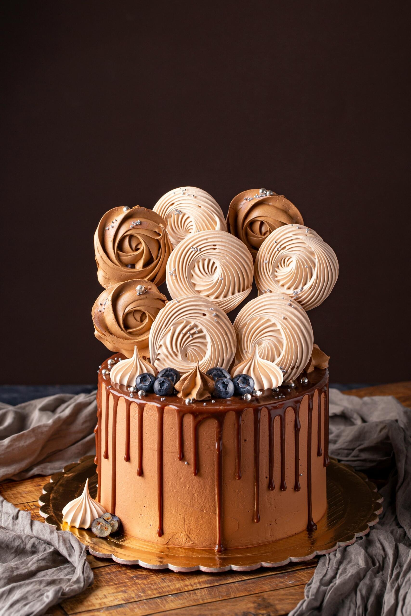 Chocolate drip cake decorated with chocolate swirls, meringues, blueberries, and silver sprinkles.