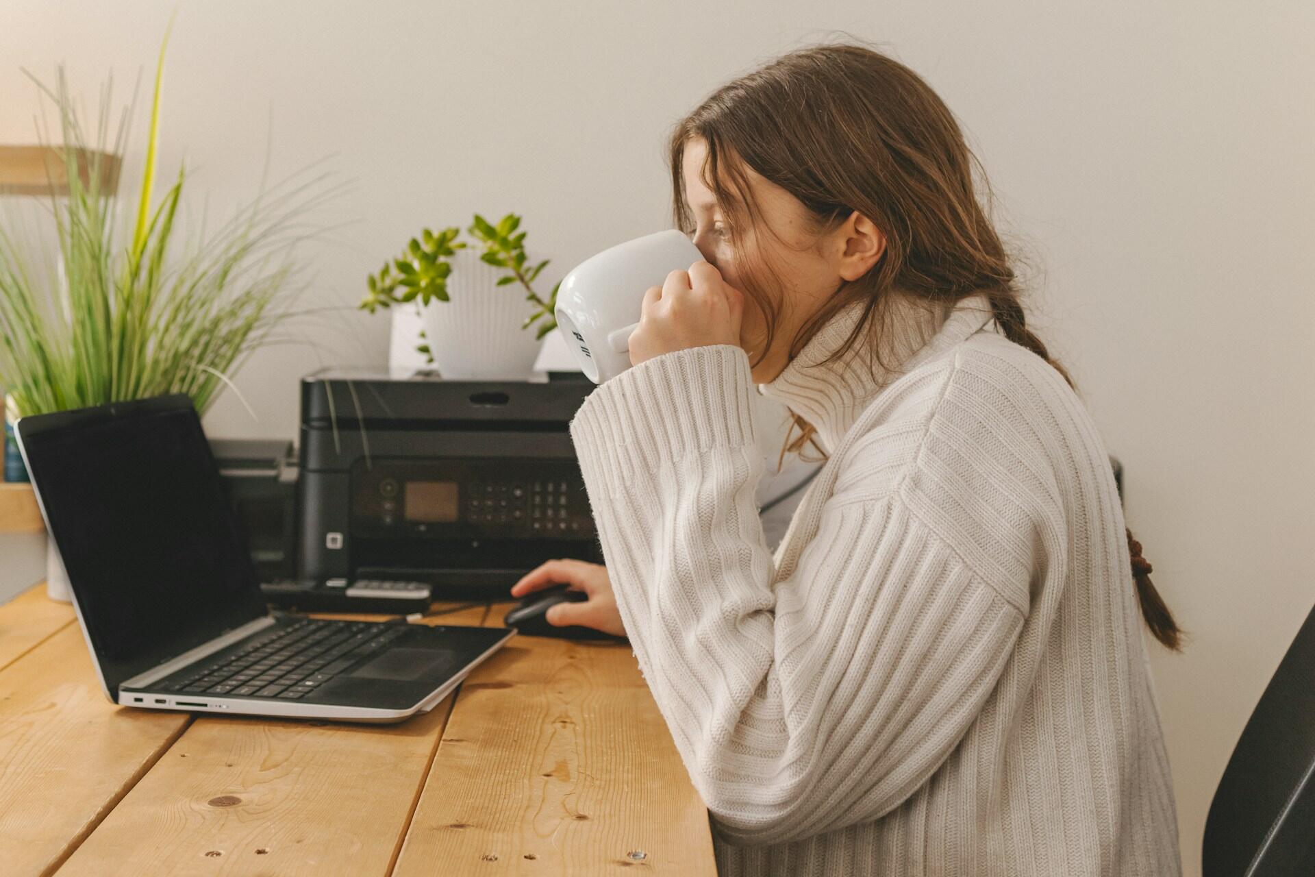 A young woman with long hair, wearing an oversized white jumper, drinks from a white mug while working on her computer.