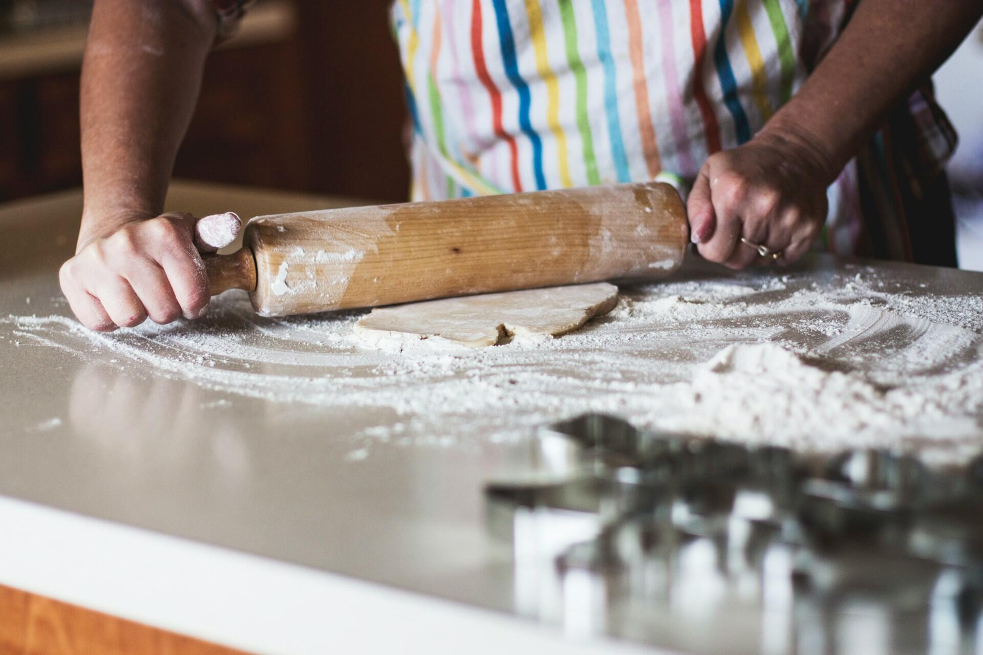 A person wearing a striped apron rolls out dough on a floured countertop with cookie cutters in the foreground.