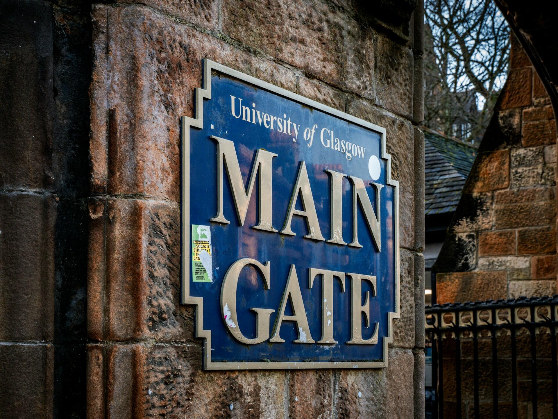 A blue rectangular sign mounted on a rustic stone pillar at the University of Glasgow reads