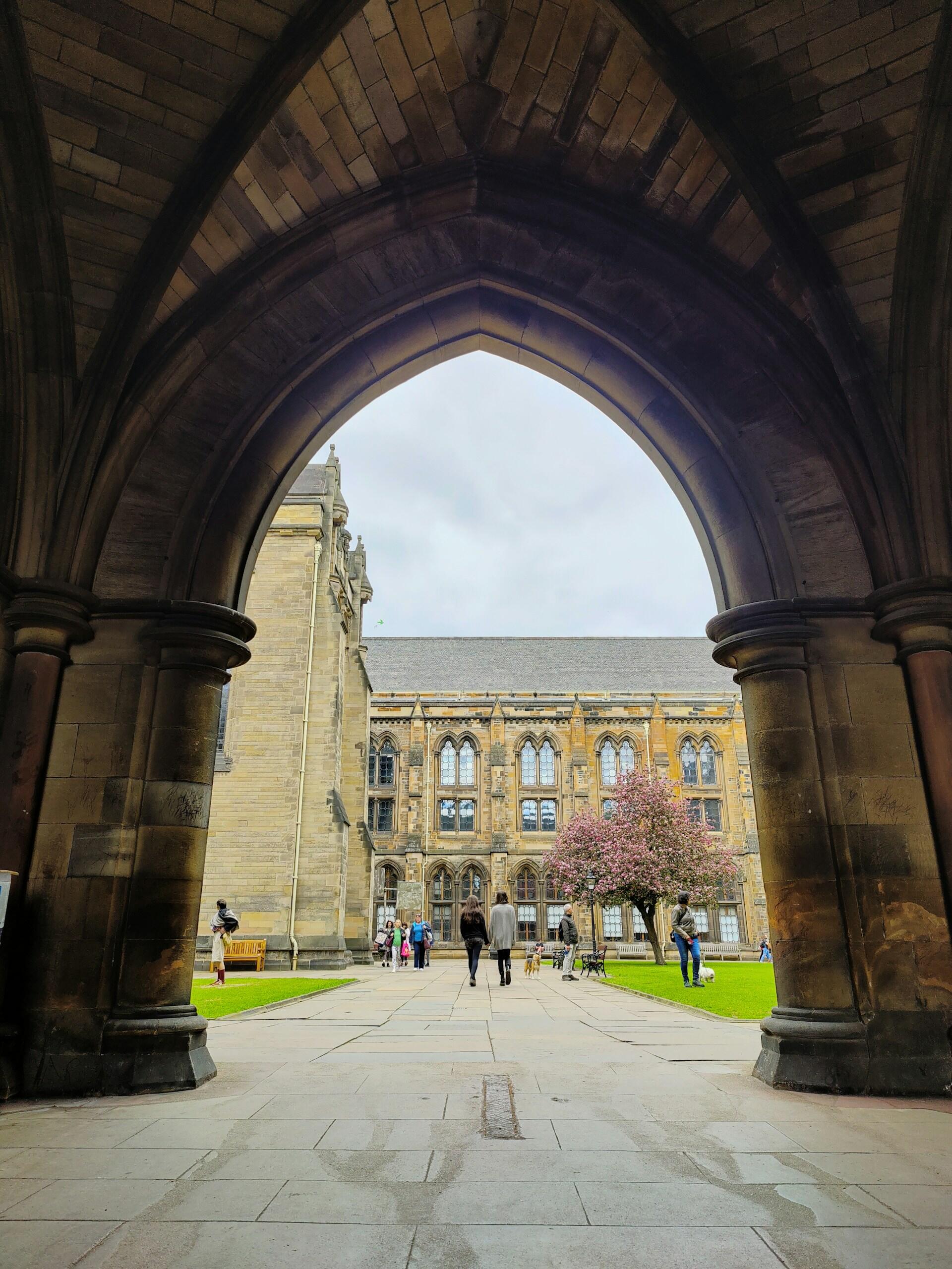 View through a large stone gothic archway looking out into a courtyard with historic buildings and people walking on a paved path.