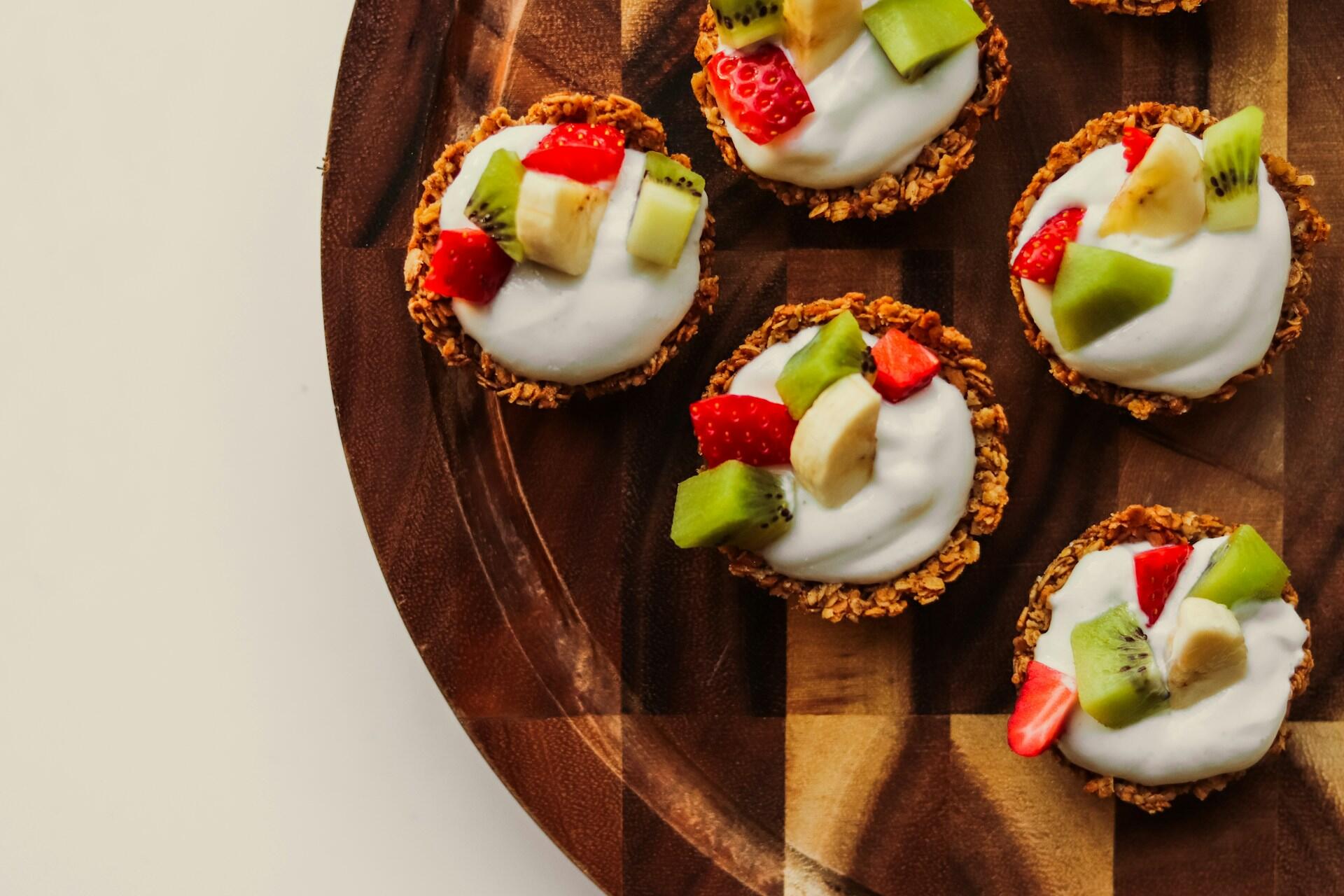 Six granola cups filled with yogurt and topped with strawberries, kiwi, and banana slices on a wooden board.