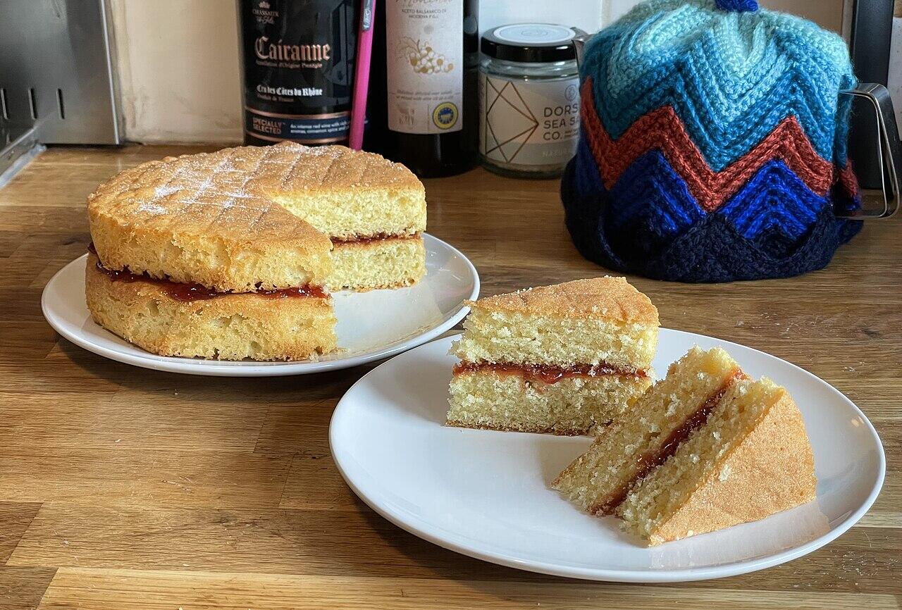 Victoria sponge cake with jam filling served on white plates with bottles and a knitted tea cosy in the background.