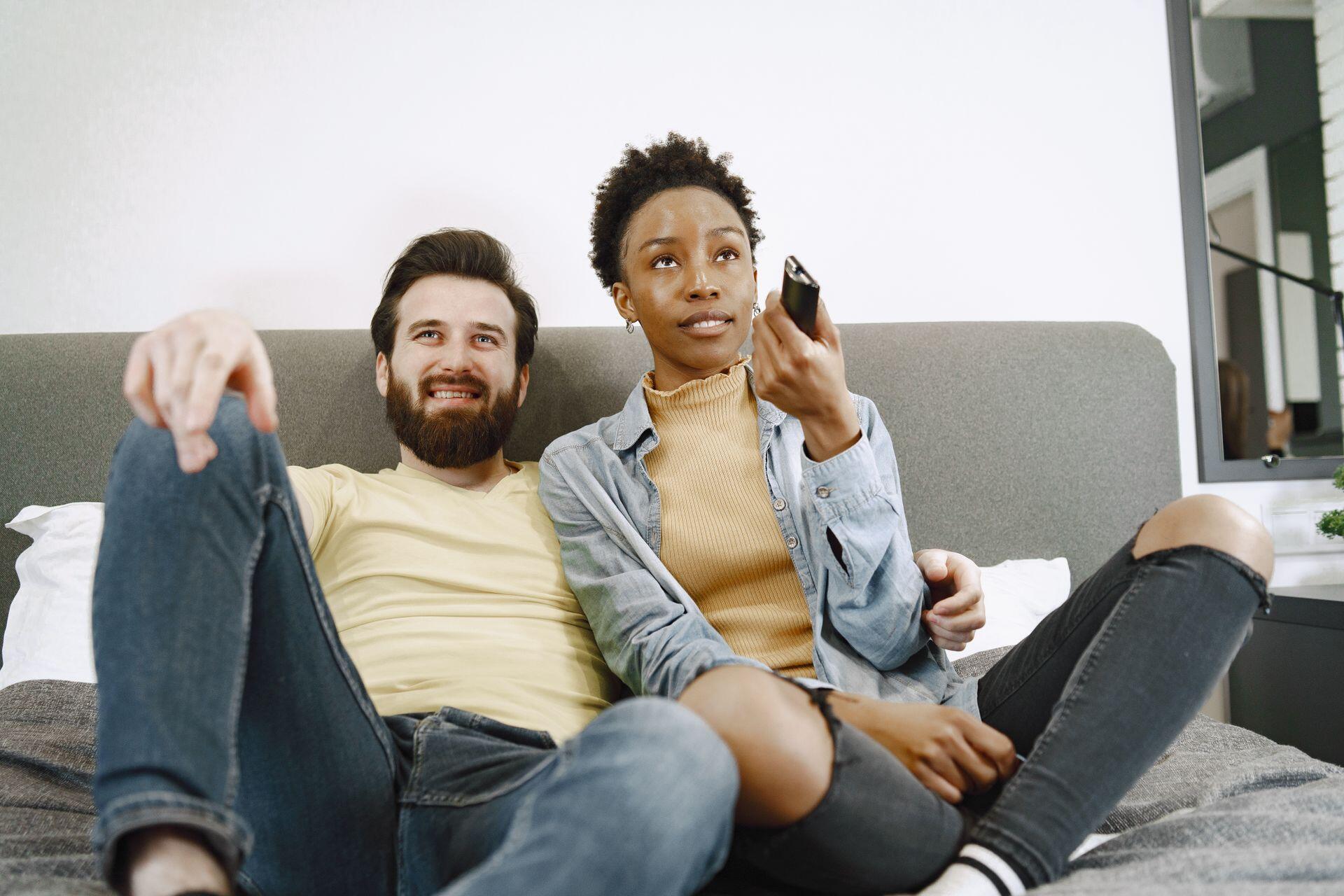 A smiling man and a woman holding a remote control sit closely on a bed, looking up.